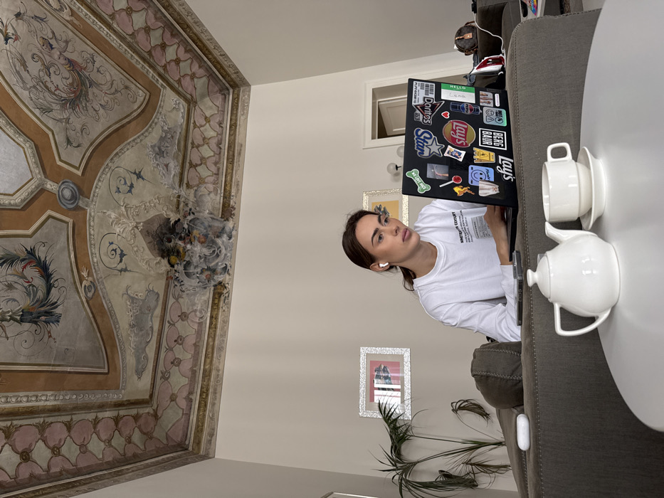 An interior scene depicts a young adult female seated on a dark grey sectional sofa, intently engaged with a black laptop covered in various stickers including brand logos like "Doritos" and "Lays." She has brown hair, wears a white long-sleeved t-shirt, and white wireless earbuds. A white ceramic teapot and a matching teacup with saucer are positioned on a white round coffee table in the foreground. The room features a highly ornate, multi-paneled painted ceiling adorned with intricate classical frescoes, including central medallions with avian motifs, scrollwork, and geometric patterns in earthy tones of brown, beige, light blue, and pink. This architectural style, particularly the ceiling, suggests a historic building, indicative of a palazzo or grand apartment in the city of Venice, Italy. The walls are plain light beige, with a potted plant on the left and two framed pictures visible. A doorway is partially visible in the background on the right. An AirPods case and a smartphone are also present on the sofa.