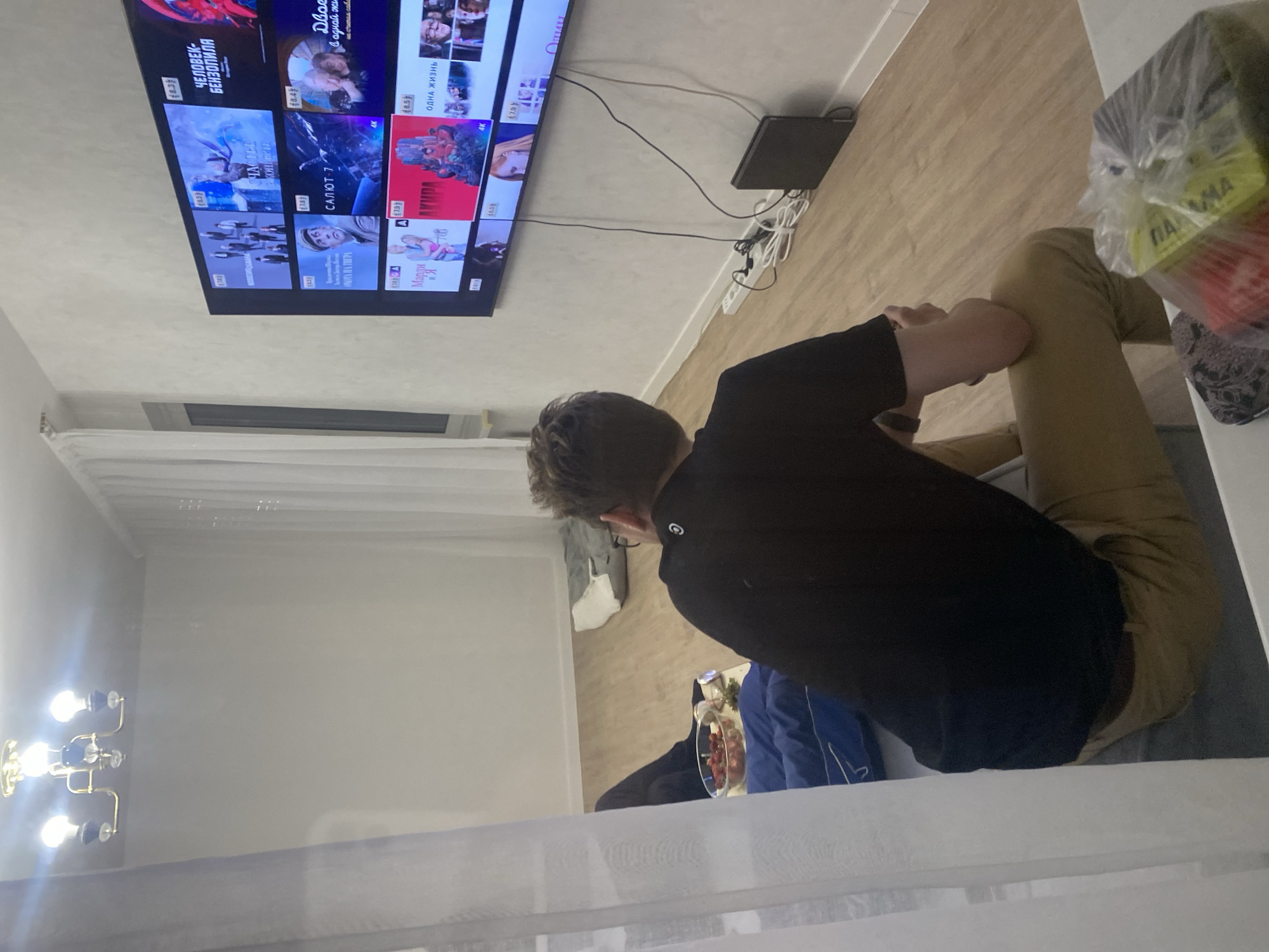The image depicts a living room scene with a large television displaying a streaming service interface. The room has light-colored walls and wooden flooring. A person, seen from behind, is seated on a couch, facing the television. They are wearing a black t-shirt and tan pants. In the foreground, a plastic bag containing some items is visible on a table. To the left, a chandelier hangs from the ceiling. The television shows various movie or show posters, with titles in Russian. There are also some cables and a black electronic device connected to the television, placed on the floor. The overall impression is a casual indoor setting. There are no visual indicators to suggest the location is specifically the city of Perm, Russia.