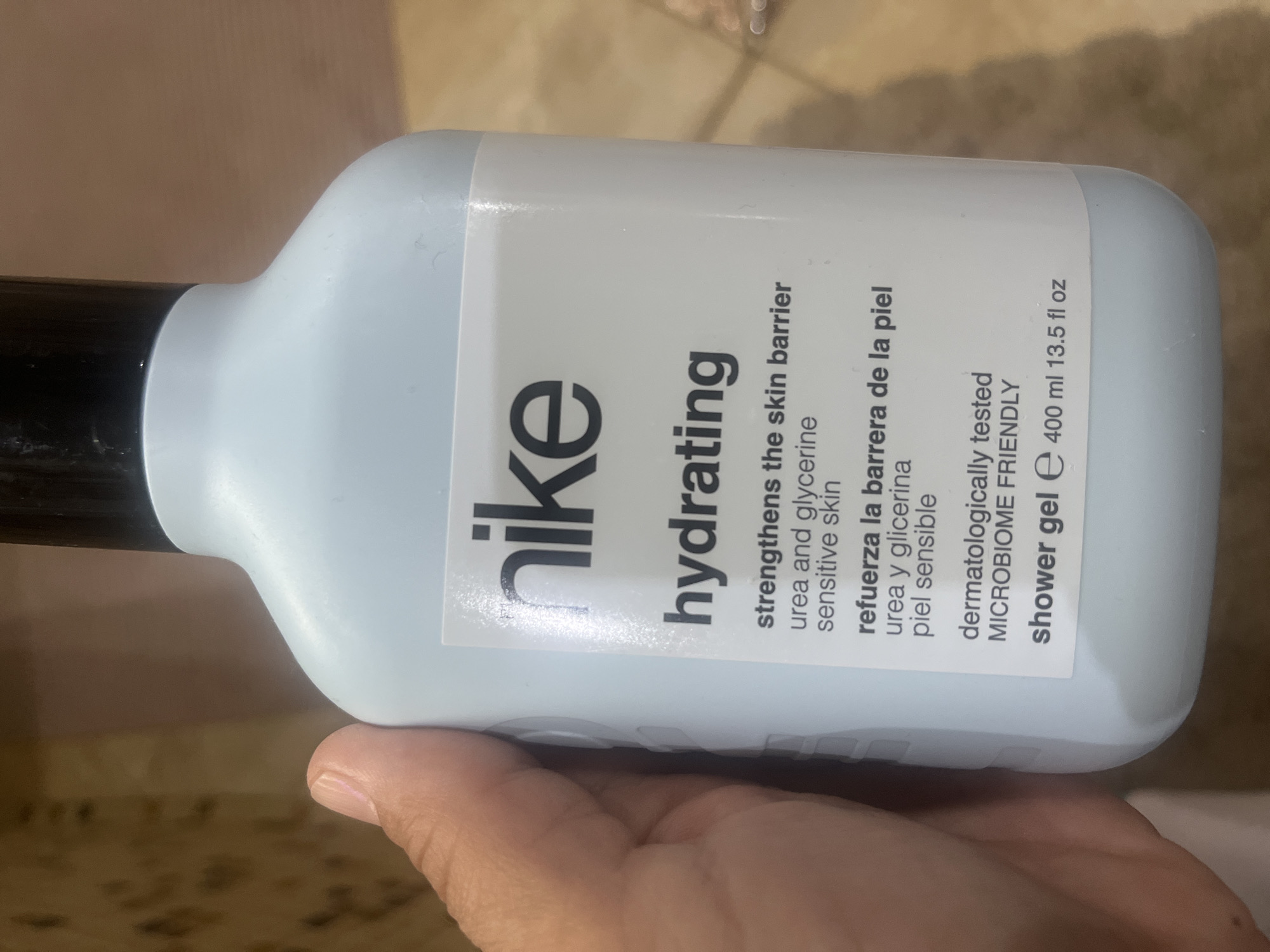 A light blue bottle of Nike Hydrating Shower Gel is held by a person's hand. The bottle has a white label with black text. The label features the Nike logo, the word "hydrating," and information about the product's benefits and ingredients in both English and Spanish. The text also indicates that the product is dermatologically tested and microbiome friendly, and states the volume as 400 ml (13.5 fl oz). The background is blurred, but appears to be a tiled or textured surface. No specific location in the city of Onokivtsi, Ukraine can be identified from the image.