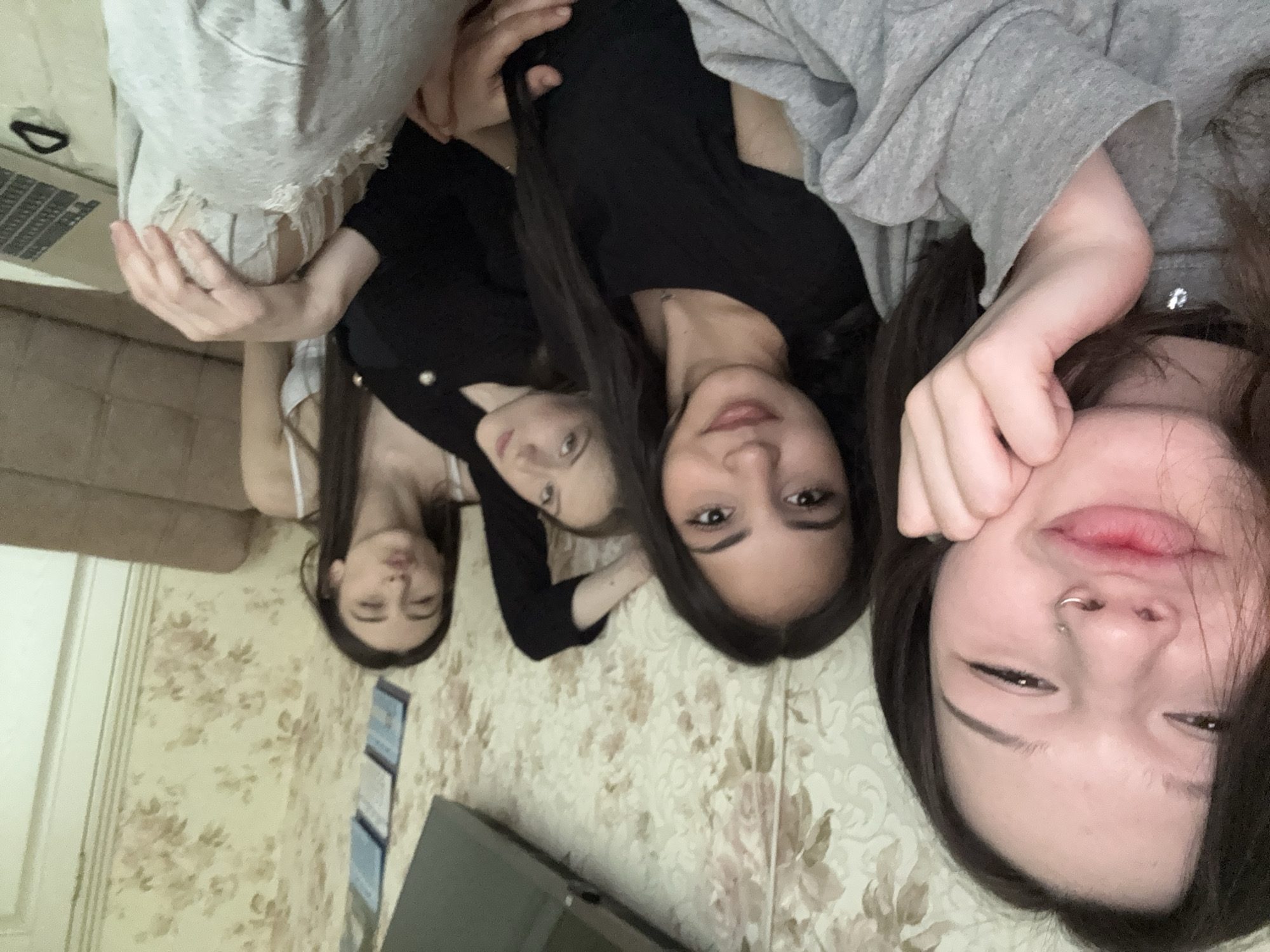 The image shows a close-up, tilted shot of four young women lying close together, possibly on a bed or floor with floral wallpaper. The women are positioned in a row, with their heads close to each other. The woman furthest to the right is closest to the camera. She has dark hair and a nose ring and is touching her cheek with her hand. The woman next to her has dark hair and a black top. The next woman also has dark hair and a black top with buttons. The final woman is wearing a white top and has long dark hair. Part of a laptop and sofa are visible in the background. There is no specific evidence to suggest the location is Saint Petersburg, Russia.