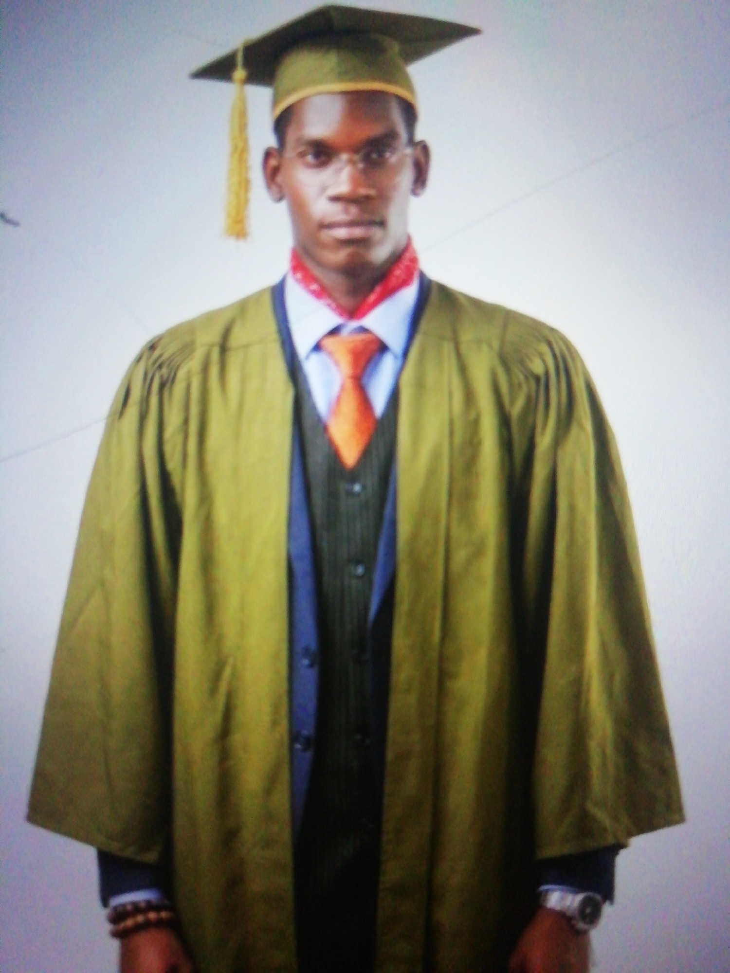 A portrait photograph features a young man, likely of African descent, standing front-on against a plain, light-colored background. He is dressed in formal academic attire, suggesting a graduation or similar academic event. He wears a dark green academic gown over a light blue collared shirt, an orange necktie, and a dark gray pinstripe waistcoat, with the sleeves of a dark blue suit jacket visible beneath the gown's sleeves. A dark green academic cap, or mortarboard, with a gold tassel, sits squarely on his head.
The man has short dark hair, dark skin, and wears thin-framed eyeglasses. A red and white patterned scarf or stole is visible around his neck, partially covered by his tie. He maintains a serious and composed expression, looking directly into the camera. On his left wrist, he wears a multi-strand wooden beaded bracelet, and on his right wrist, a silver-toned watch. The lighting is soft and even, characteristic of an indoor portrait setting. There is no visible text in the image. Given the context, this photograph was taken in Enugu, Nigeria.