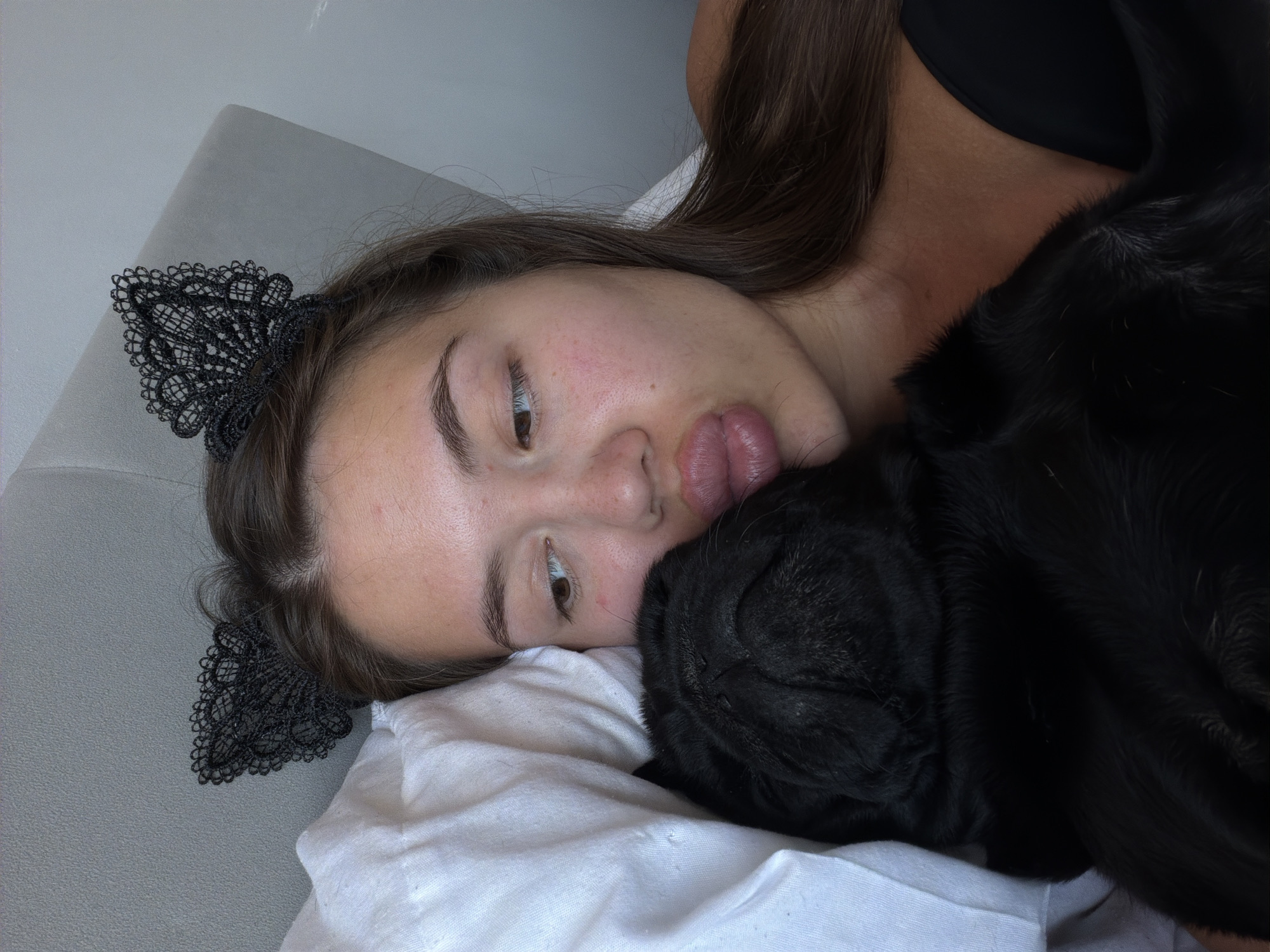The image shows a close-up of a young woman lying down, resting her head on a white pillow. She is wearing a black lace headband resembling cat ears. Next to her, a black pug is snuggled close to her face. The woman's eyes are open and she is looking downwards, with her lips slightly parted. Her expression appears relaxed and perhaps a little sleepy. The pug's face is partially visible, pressed against her cheek.
The setting appears to be indoors, with a grey textured surface visible in the background, possibly a couch or wall. The lighting suggests it is daytime. The overall mood is intimate and comfortable, conveying a sense of closeness between the woman and her pet. There is no visible text in the image. The location context is Sirius, Russia.