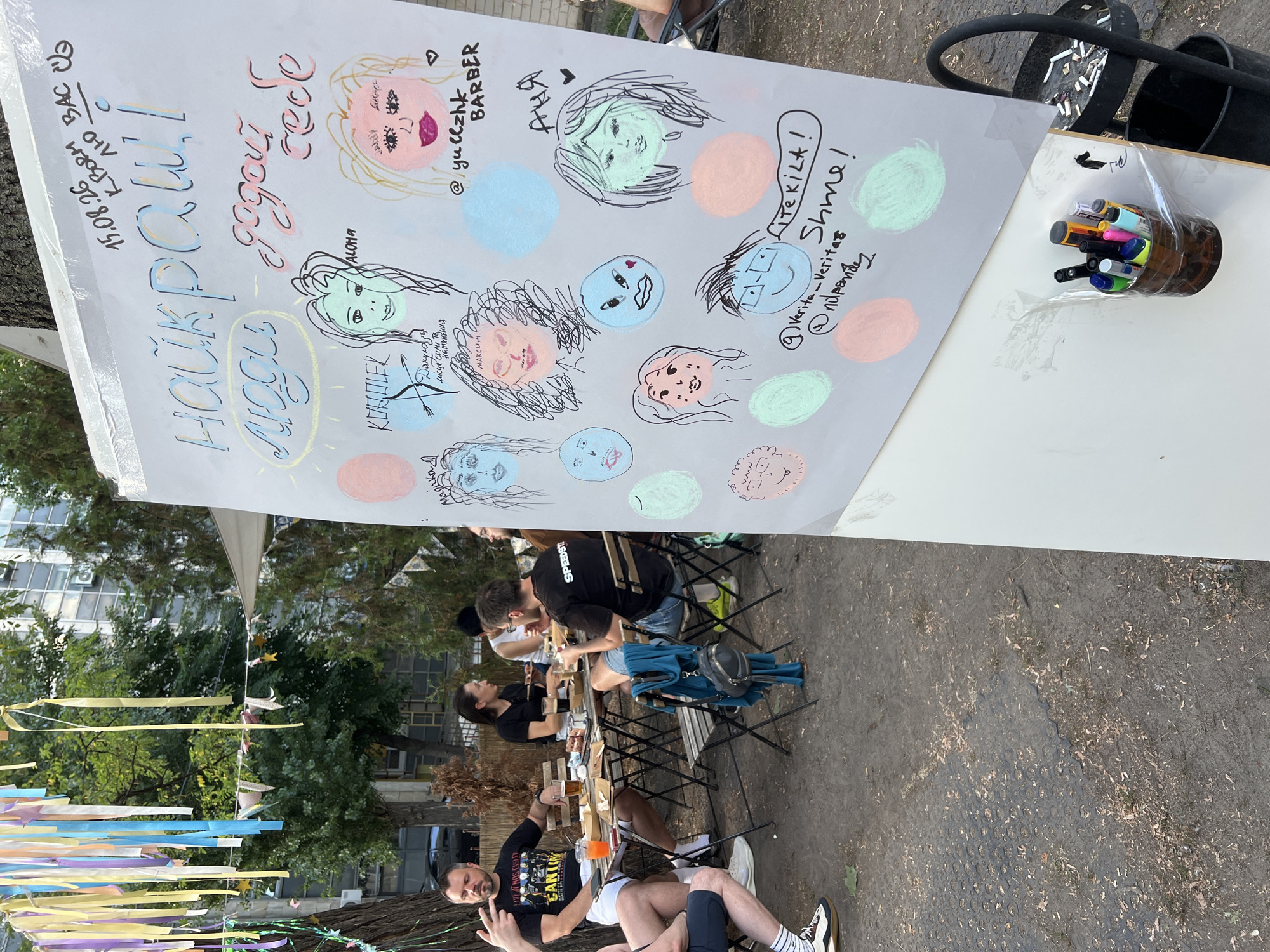 The image captures an outdoor gathering, likely a street art or community event, in Kyiv, Ukraine. The focal point is a large whiteboard covered in chalk drawings and handwritten text. The drawings depict various stylized portraits of people, with colorful circles drawn around some of them. Prominently displayed at the top of the whiteboard is the date "15.08.26" followed by Ukrainian text that translates to "best wishes to you."
In the background, several people are seated around tables, appearing to be enjoying food and drinks. They are dressed casually, and the atmosphere suggests a relaxed social gathering. The environment includes trees and buildings, typical of an urban setting. To the right of the whiteboard, there's a receptacle filled with cigarette butts, indicating a smoking area. A collection of markers and pens is attached to the base of the whiteboard, ready for use. The overall scene suggests a vibrant, community-oriented event taking place outdoors in Kyiv.