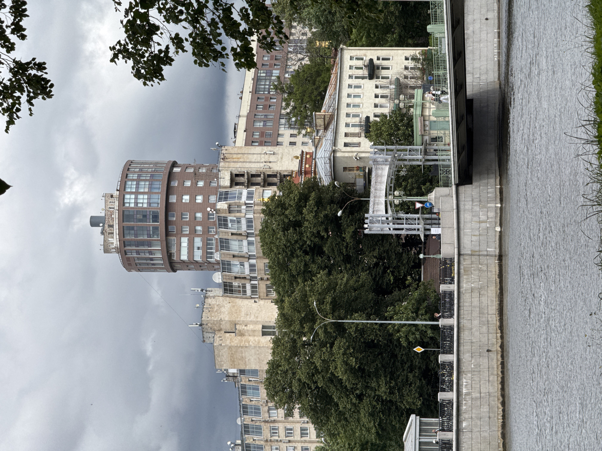 This is a photograph of a cloudy day in Saint Petersburg, Russia, likely taken from across a body of water. In the foreground, a grey, rippling river occupies the lower half of the image. A substantial concrete embankment with a decorative railing runs along the riverbank. Beyond the embankment, a bridge spans the river, with its concrete supports visible. On the bridge, several individuals can be seen walking. A light green kiosk or small building is positioned on the bridge, adjacent to a lamppost.
Behind the embankment and bridge, a densely wooded area of green trees provides a natural backdrop. Above the trees, a cluster of multi-story buildings dominates the cityscape. The most prominent structure is a distinctive, circular, reddish-brown building with numerous windows, situated towards the center of the frame. To its left and right, other buildings of varying architectural styles and heights are visible, some appearing to be older, with beige or light-colored facades, and others are modern apartment buildings with many windows. The sky is overcast with heavy, grey clouds, suggesting a potentially cool or rainy day.