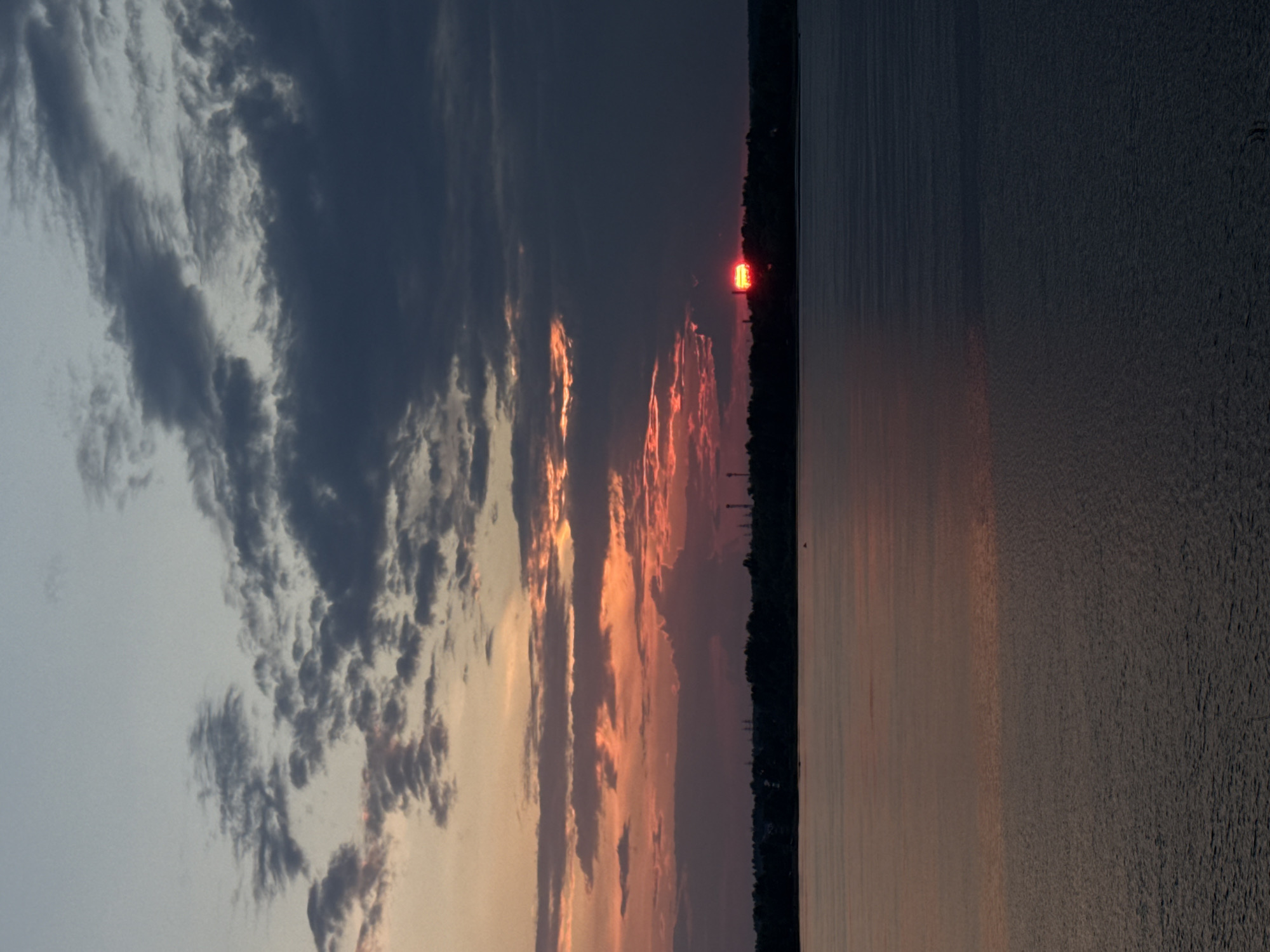 The image depicts a sunset over the Kama River in Chaikovsky, Russia. The sky is filled with dramatic clouds, ranging from pale blue at the top to dark gray and vivid orange and red hues near the horizon. The sun is a bright red orb, partially obscured by clouds, setting behind a dark silhouette of trees and industrial structures on the distant shore. The river reflects the colors of the sky, creating a muted, rippling surface that transitions from warmer tones in the foreground to darker blues in the distance. The overall scene is one of natural beauty with a touch of industrial presence.
