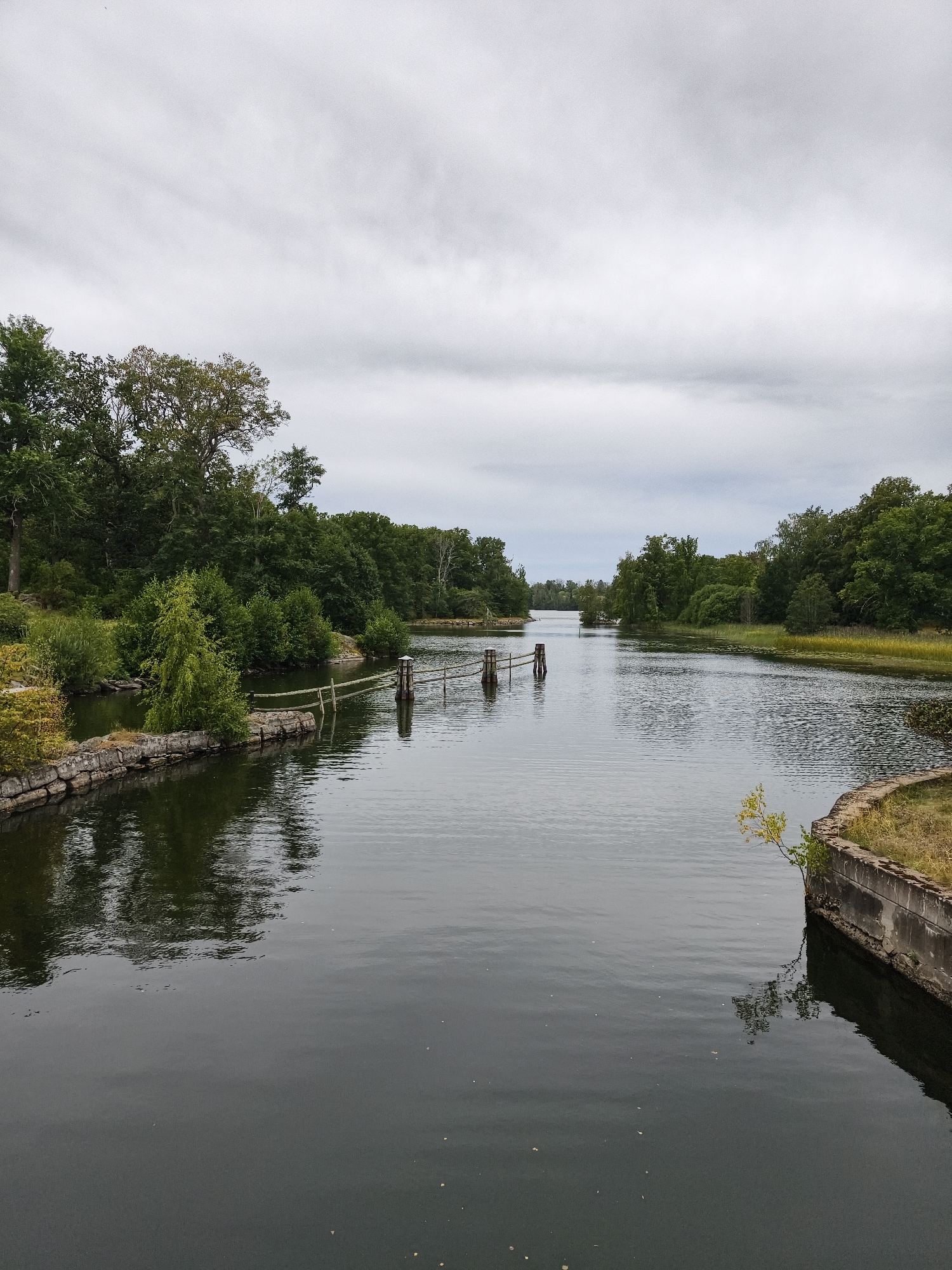 The image depicts a wide, dark canal or river flowing through a wooded area, likely in Brokind, Sweden. The water is calm and reflects the overcast sky and surrounding trees. The left bank of the canal is lined with rocks and vegetation, with a dilapidated wooden fence extending into the water. The right bank features a concrete structure, also with vegetation growing on and around it. Dense green trees dominate both sides of the canal, creating a lush, natural landscape. The sky is filled with thick, grey clouds. There are no people visible in the scene, and no apparent actions are taking place.