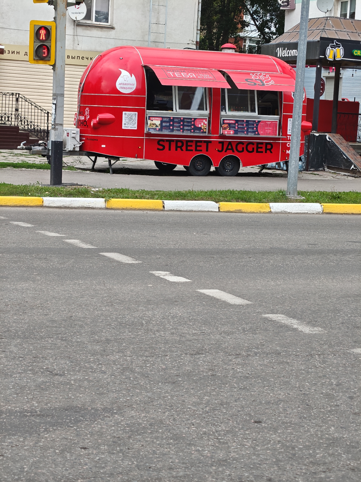 A red food truck, named "STREET JAGGER," is parked on a paved street in Petropavl, Kazakhstan. The food truck has a retractable awning and menu displays with food items. A traffic light with red and green signals is visible to the left of the food truck, along with a utility pole and some buildings in the background. The street is marked with white dashed lines. The curb is painted with alternating yellow and white stripes. The scene is captured from a slightly elevated perspective, looking down at the street and the food truck.
