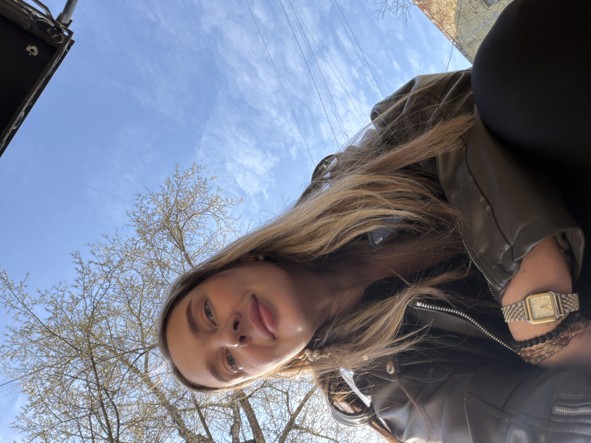 A woman takes a selfie from below, revealing a clear sky with thin clouds, trees, and a street. The woman has long blonde hair and wears a leather jacket, a watch, and bracelets. The location appears to be Moscow, Russia, based on the architecture and cityscape visible in the background. Power lines stretch across the sky, and a dark object, possibly part of a building or street fixture, is visible in the upper left corner.