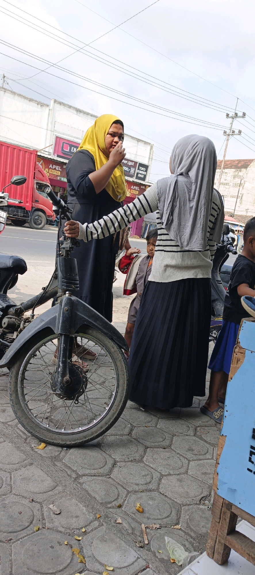The image depicts an outdoor scene on a sidewalk, likely in Kasiyan, Indonesia, with a view of a street and buildings in the background. In the foreground, a motorcycle is partially visible, with its front wheel and fender prominently featured. Two women wearing hijabs are the main subjects. The woman on the left, wearing a yellow hijab and a black dress, is seen holding her hand to her mouth, possibly eating or speaking. She is standing next to the motorcycle. To her right, another woman in a grey hijab and a striped long-sleeved shirt over a black pleated skirt stands facing away from the camera, with her right arm extended towards the woman in yellow. A young child, wearing a brown jacket over an orange shirt, is visible behind the woman in the grey hijab. A glimpse of another child in a black t-shirt and blue shorts is seen further to the right. The environment includes a paved sidewalk with hexagonal stones, a red truck in the background, and a building with signage that includes the word "APEL". There are scattered leaves and debris on the ground. A blue wooden structure is present on the far right of the image. The sky is overcast with visible power lines.