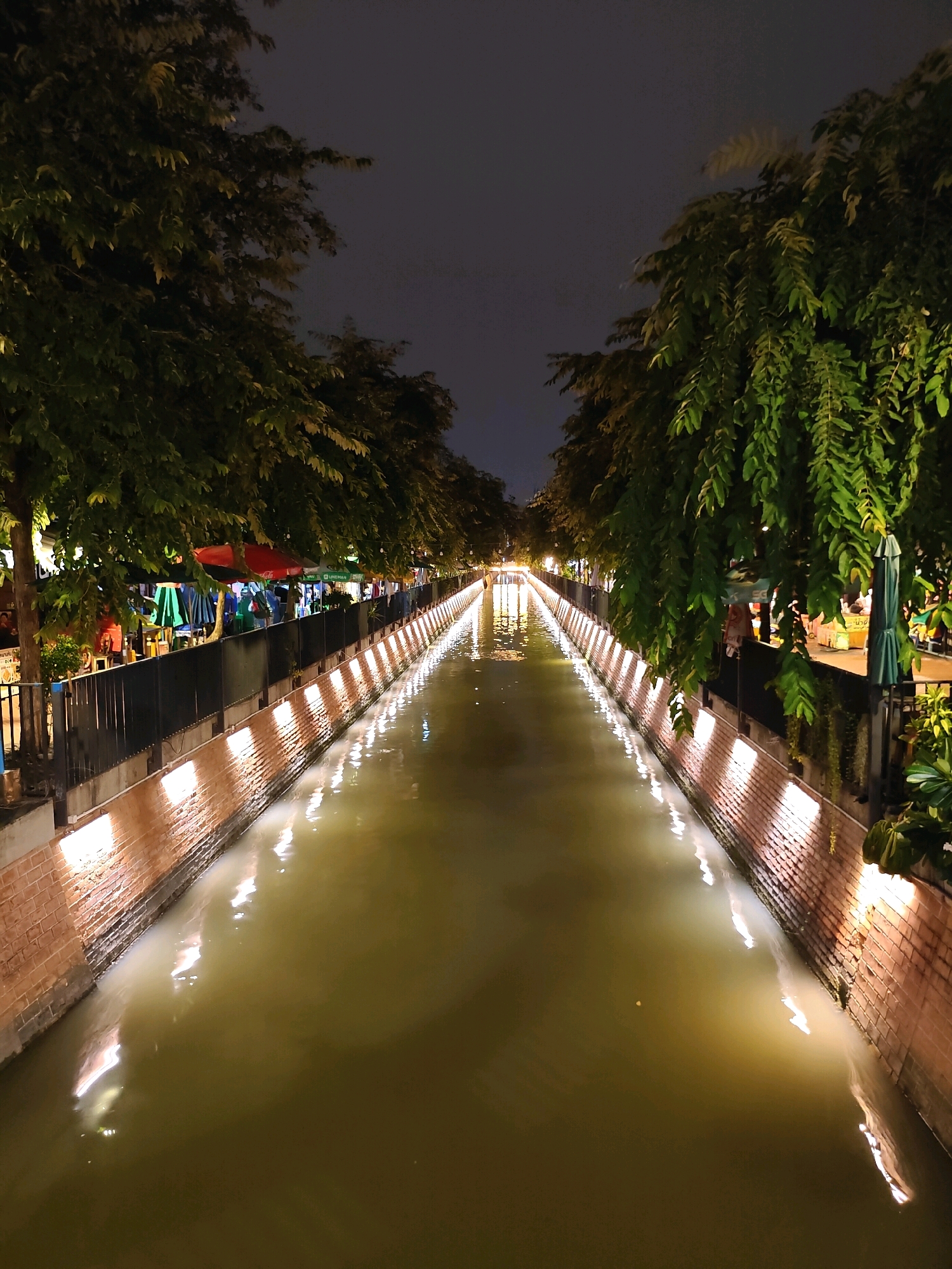 The image shows a canal at night in Bangkok, Thailand, likely at a market or dining area, possibly the Talad Neon Night Market or a similar riverside establishment. The canal is lined with brick walls and illuminated by a series of lights that reflect on the water's surface, creating a warm glow. Lush green trees with long, trailing leaves hang over the canal on both sides, casting shadows and adding to the ambiance. Along the banks, there are market stalls and seating areas with umbrellas, suggesting a place for dining and shopping. Several people are visible, scattered around the stalls and walking along the walkway. The overall atmosphere is lively and illuminated, a typical scene of a vibrant night market in Bangkok.