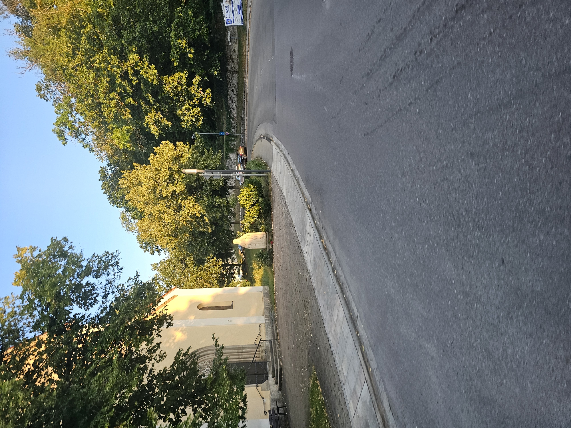 The image depicts a street scene in Pappenheim, Germany, likely during the late afternoon or early evening due to the warm sunlight illuminating the trees and building. On the left side of the frame is a light-yellow building, possibly a church or public institution, with a dark arched doorway and a single arched window. In front of the building, there is a paved walkway with a dark bench. Adjacent to the walkway is a statue of a robed figure. The street itself is paved and curves to the right, with a sidewalk separating it from the walkway. Lush green trees line the upper edge of the scene, casting shadows on the road and buildings. In the distance, a stone wall runs along a wooded hillside. A few cars are visible on the road, including one white car and a darker sedan. A blue parking sign is also present. A large white billboard with German text, including "170 JAHRE", is situated in the background on the right side of the road. The sky is clear and blue.