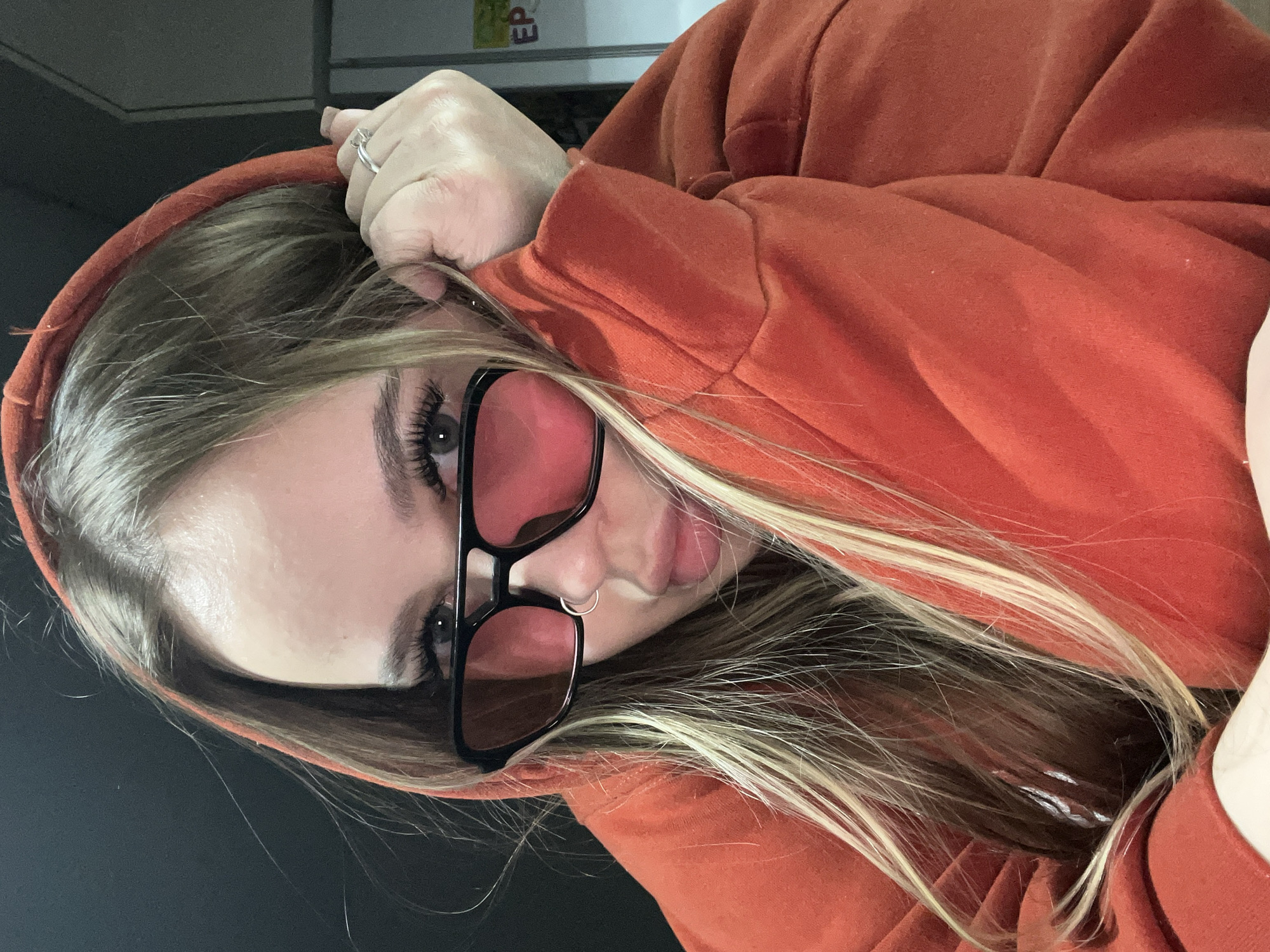A woman with blonde hair, wearing a reddish-orange hooded sweatshirt and black-framed sunglasses with red lenses, is the primary subject of the image. She is wearing a nose ring in her left nostril and a ring on her left hand. The background is slightly out of focus and dark, with a partially visible white box in the upper left. The image appears to be taken indoors. The location and its affiliation with Krasnodar, Russia, cannot be determined from the image.