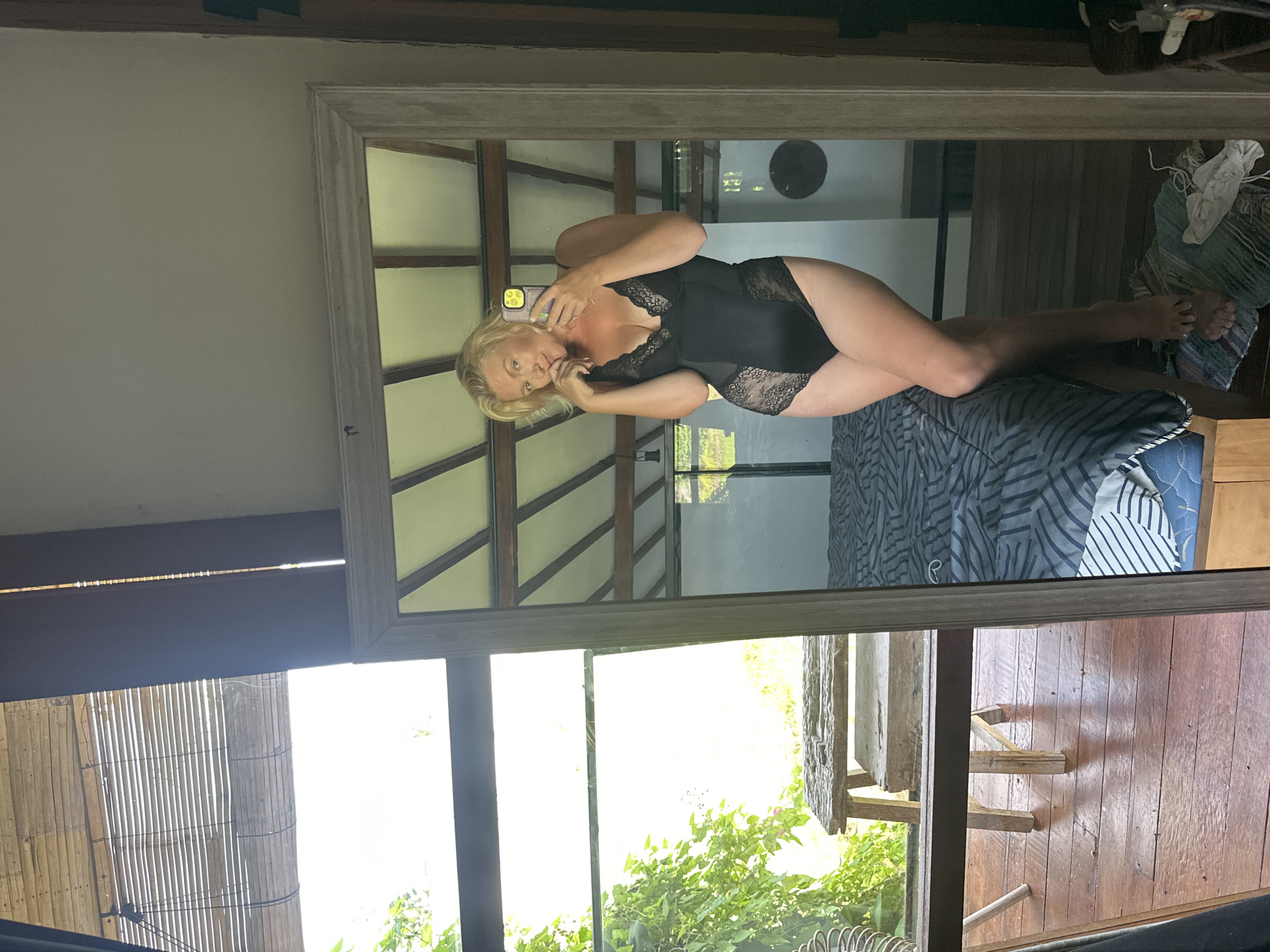 A blonde woman in a black lace bodysuit poses for a mirror selfie in a room with a slatted bamboo blind covering a window, a wooden floor, and a bed with striped bedding. A wooden bench is visible outside the window. The reflection in the mirror shows the woman holding a phone and touching her chin. The room appears to be in a villa or resort in North Kuta, Indonesia.