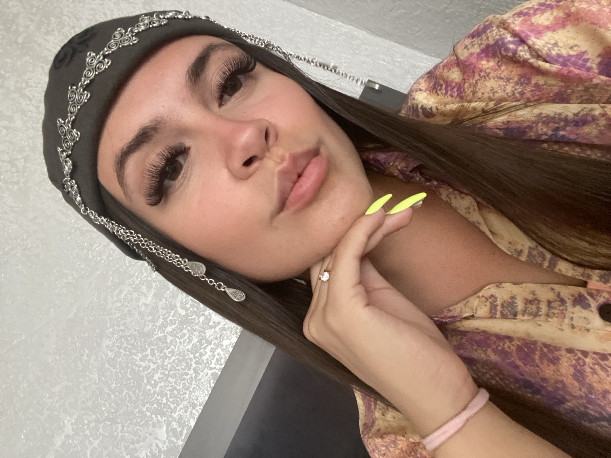 A young woman with long, dark hair is shown from the chest up, looking towards the camera with her chin resting on her hand. She is wearing a dark beanie adorned with silver chain detailing and teardrop-shaped charms. Her fingernails are painted a bright neon yellow. She is also wearing a patterned shirt with shades of purple, pink, and yellow. A thin pink bracelet is visible on her wrist, and she has a ring on her right ring finger. The background appears to be an indoor setting with a textured white wall. There is no information in the image to suggest it is located in Chalda, Russia.