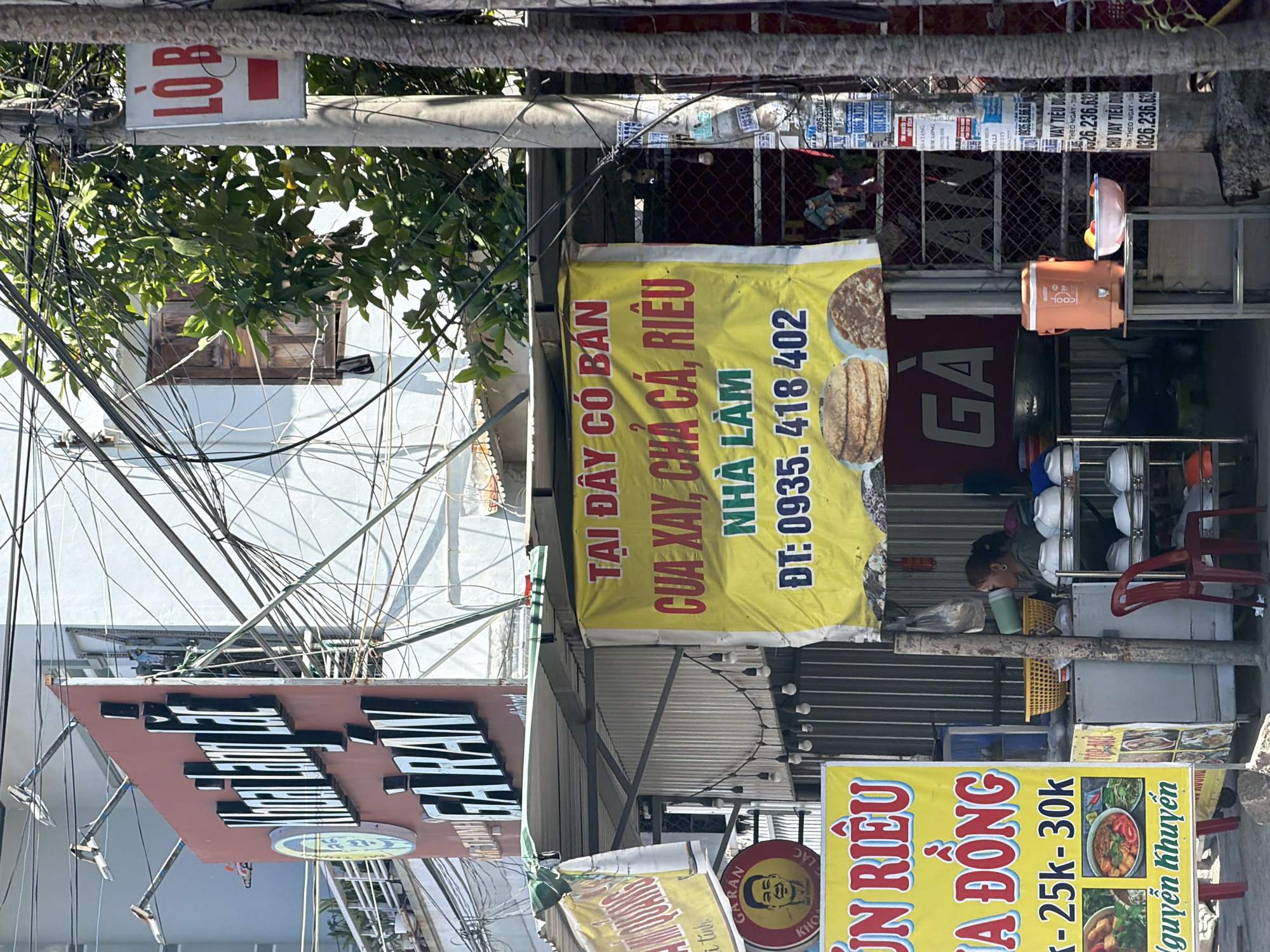 The image shows a street scene in Nha Trang Ward, Vietnam. A food stall with a large yellow banner advertising "CUA XAY, CHA CA, RIEU" (crab, fish cake, and crab paste noodles) is prominently displayed. A woman is seen behind the counter, serving food. The stall is decorated with various signs and images of food. In the background, there are buildings, power lines, and trees. The overall impression is of a busy urban environment with local commerce.
