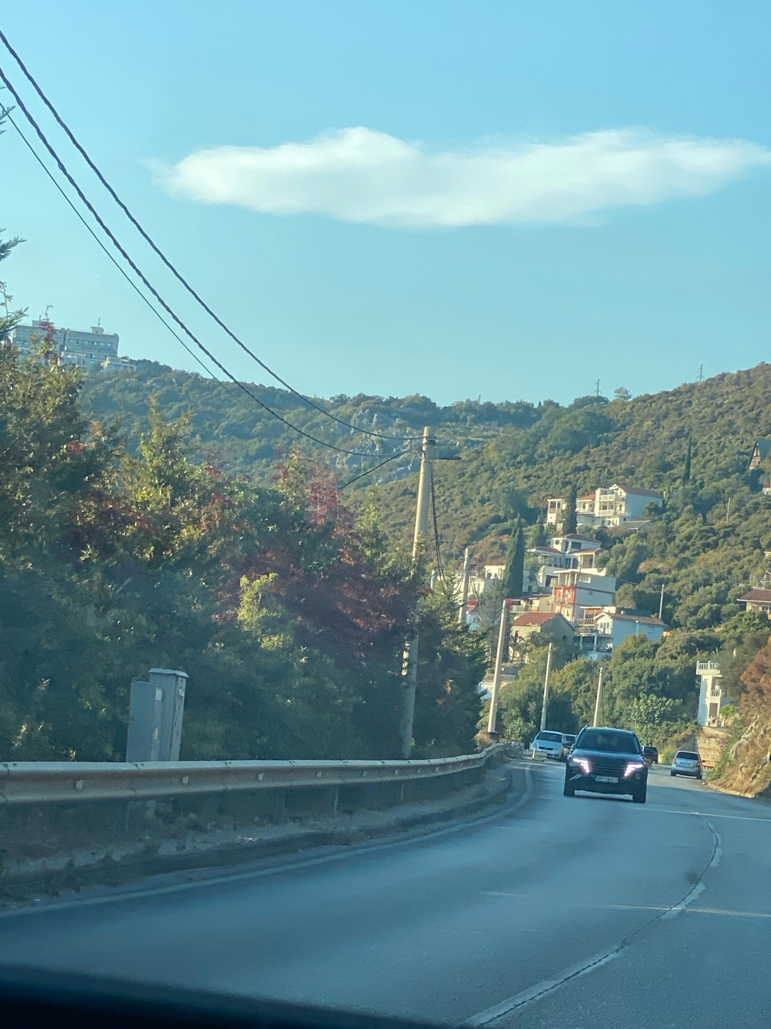 The image shows a winding road in Dobra Voda, Montenegro, with houses scattered on a lush, green hillside. Several vehicles are driving on the road, including a black SUV in the foreground. Overhead power lines crisscross the sky, and a single, elongated cloud is visible against the clear blue sky. The road is bordered by a metal guardrail on the left, and a steep, vegetated slope rises on the right. The scene appears to be captured during daylight, with the sun casting light on the landscape.