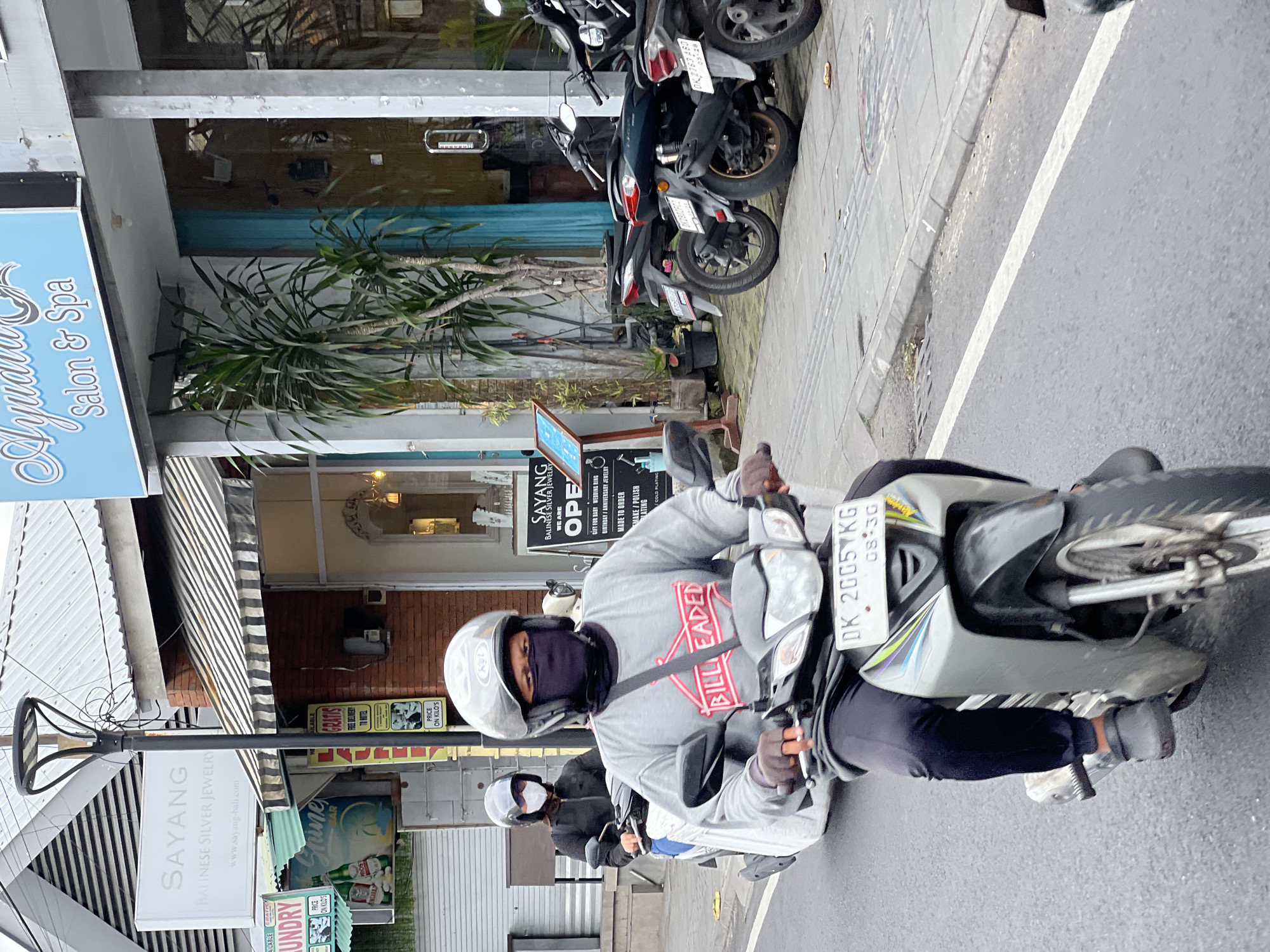 A man wearing a helmet and face mask rides a scooter on a street in Denpasar, Indonesia. He is wearing a grey sweatshirt with the words "BILL LEADED" printed in red. Behind him, a woman also wearing a helmet and face mask rides another scooter. They are both on a road with shops and parked motorcycles on the side. The shops have signs for "Sayang Balinese Silver Jewelry" and "Ayudia Salon & Spa." The weather appears overcast.