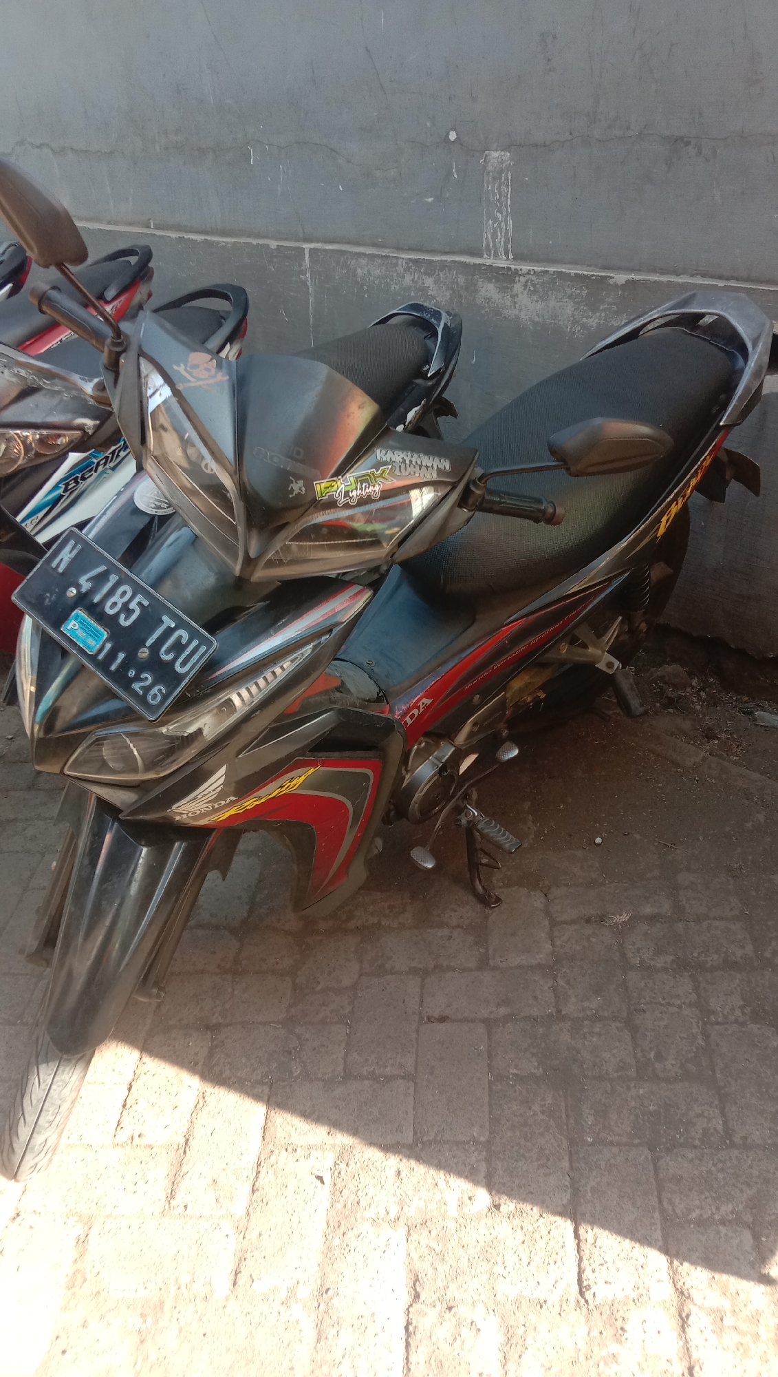 The image shows two black motorcycles parked side-by-side on a paved surface. The motorcycle in the foreground is a black Honda with red and yellow decals on its side panel and front. It has a license plate with the number N 4185 TCU and the text "11.26". A sticker with a skull and crossbones and "FK Lighting" is visible on the front fairing. The second motorcycle, partially obscured, appears to be a similar model and color. They are parked against a textured gray concrete wall. The ground is paved with rectangular bricks and a diagonal shadow from an unseen object falls across the lower portion of the image. The scene appears to be an outdoor area in Karanganyar, Indonesia, likely a street or parking lot, given the license plate format and the presence of motorcycles.