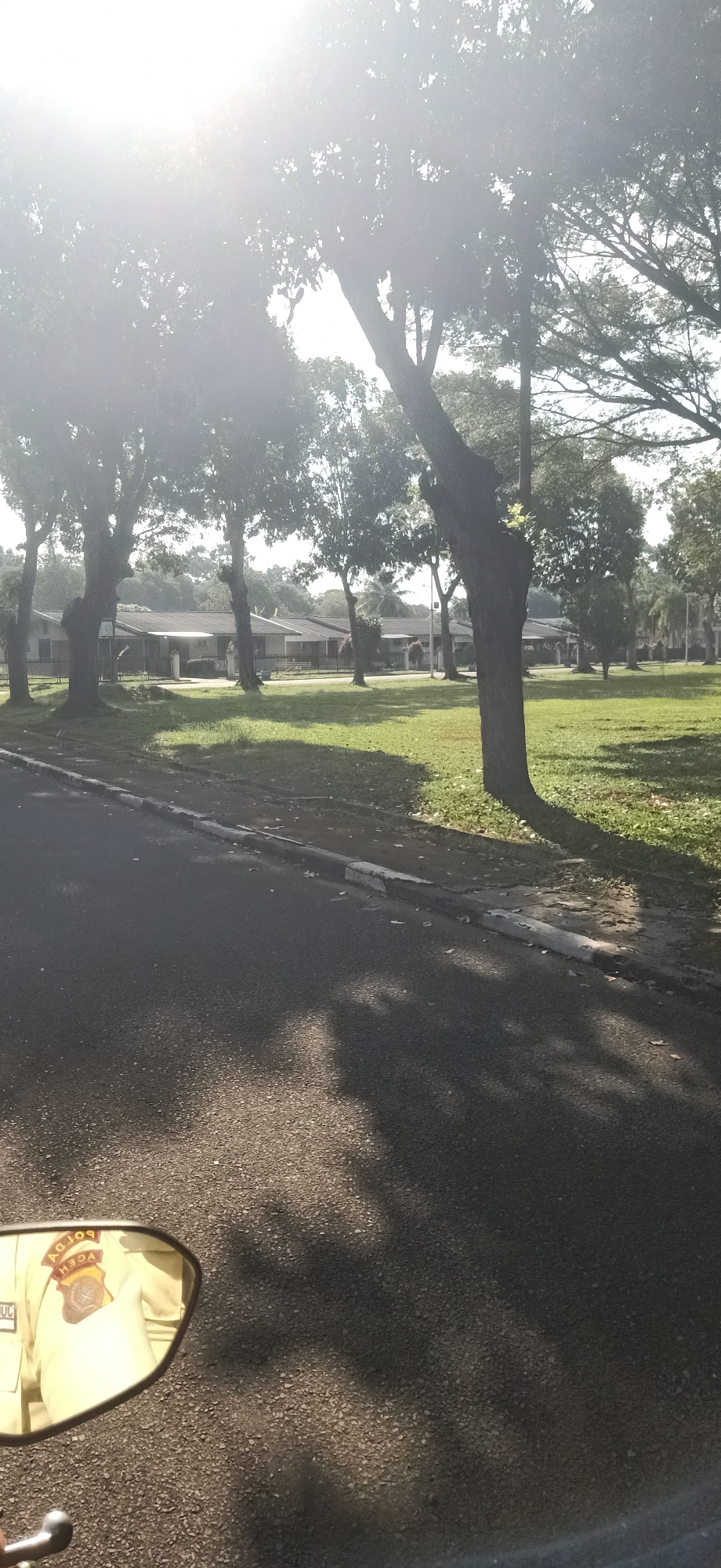 The image depicts a view from a vehicle, likely a motorcycle, looking out onto a street and a grassy area with trees. In the foreground, the asphalt road is visible, with shadows cast by the trees. To the right, there is a curb and a grassy area with scattered leaves. Beyond the grass, a line of single-story buildings with tiled roofs is visible, partially obscured by trees. The buildings appear to be residential or institutional in nature. The sky is bright and appears to be sunny, with some lens flare from the sun. In the lower left corner of the image, a motorcycle's rearview mirror is visible, reflecting a person wearing a light-colored uniform with insignia. The insignia in the mirror includes the word "POLDA" and "ACEH," suggesting the person is a police officer in Aceh province, Indonesia. The overall impression is an outdoor scene, possibly in a suburban or semi-rural area within Palohgadeng, Indonesia, during the daytime.