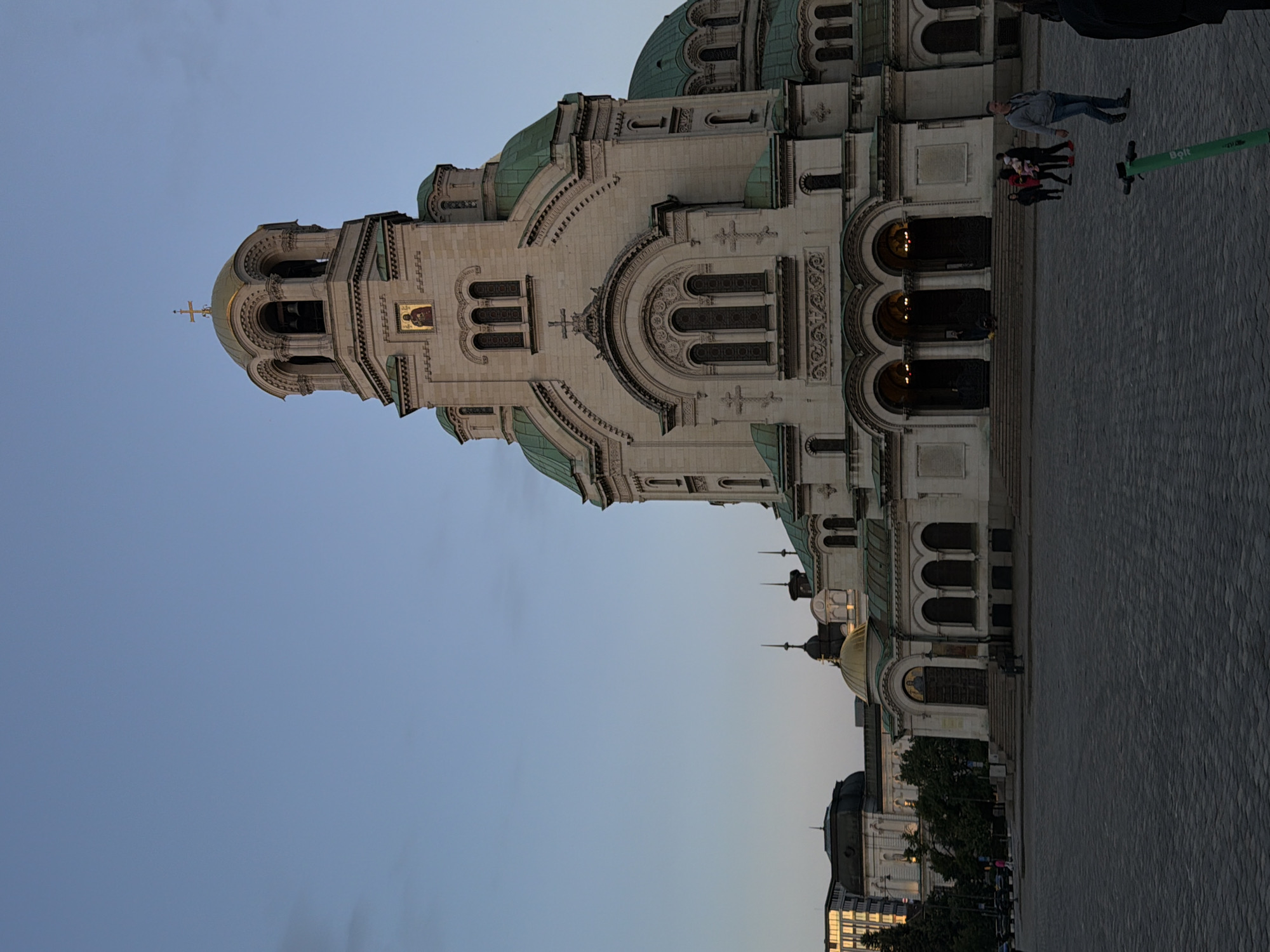 The image depicts the Alexander Nevsky Cathedral in Sofia, Bulgaria. The cathedral is an ornate, neo-Byzantine structure with multiple domes covered in green patina. The building's façade is white stone, richly detailed with arches, windows, and decorative elements. A golden-domed tower with a cross tops the cathedral. The cathedral sits on a paved surface. In the foreground, a group of people are walking, and a green electric scooter is visible. The sky is a clear, light blue. Part of the cityscape is also visible near the bottom.
