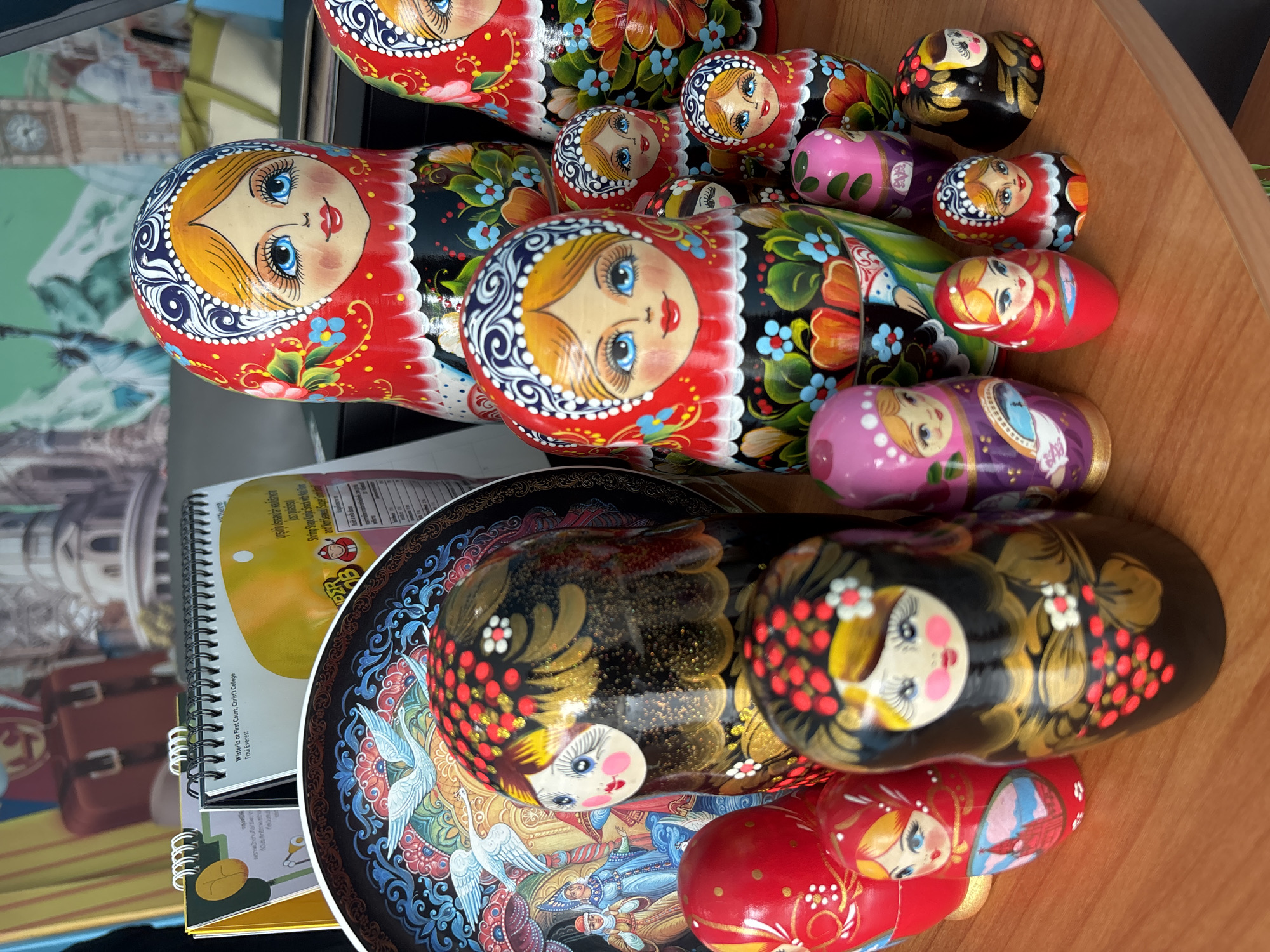 The image shows a collection of Russian nesting dolls, also known as Matryoshka dolls, arranged on a surface that appears to be a wooden desk or table. The dolls are of various sizes, with the largest at the front and progressively smaller dolls behind. They feature traditional Russian folk art designs, including floral patterns, red and black color schemes, and stylized facial features. One set rests on a decorative plate with a blue and gold painted scene of people and birds. To the left of the plate, there's a notebook or book with a spiral binding and a yellow cover. There is no verifiable evidence to indicate that the image was taken in Baan Mae Nam, Thailand. The provided information is limited to the immediate objects and environment visible in the photograph.