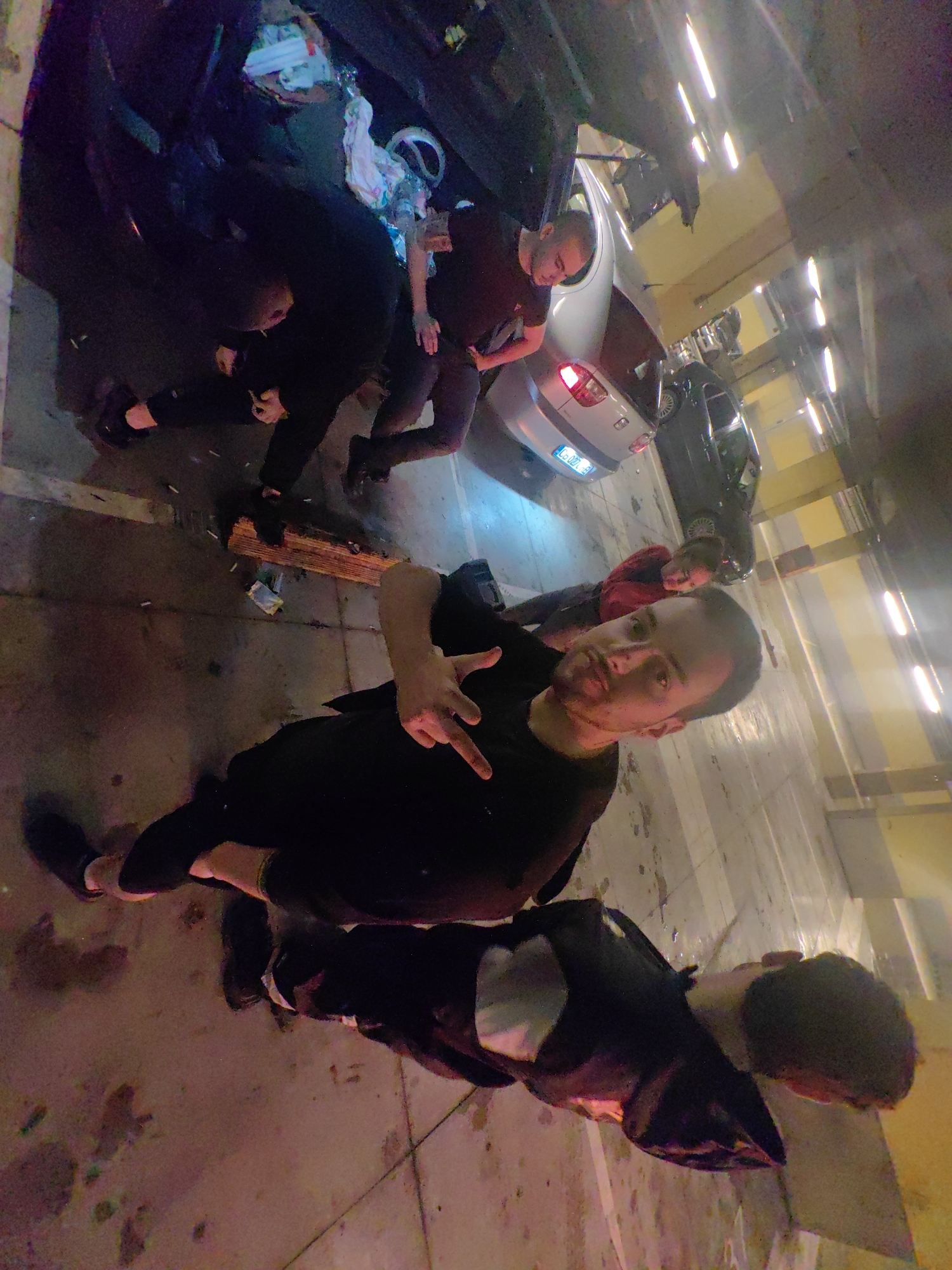 The image depicts a group of people in an indoor parking garage in Sofia, Bulgaria. The ground is concrete and marked with parking space lines. There are several cars parked in the background. The group appears to be interacting, with the person in the foreground posing for the camera. Cigarette butts and other trash are scattered on the ground near a wooden plank. The lighting is artificial and somewhat dim.