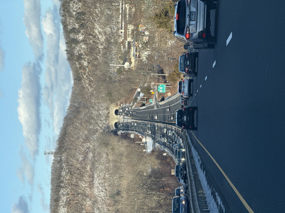 The image captures a multi-lane highway experiencing traffic congestion during daylight hours on a winter day. The scene is viewed from inside a vehicle, looking ahead down the road.
In the immediate foreground, a silver SUV is prominent in the right lane, with its rear displaying a license plate that reads "111-WYL". A black sedan follows directly behind it, and other vehicles are visible further back in the same lane. To the left, an adjacent lane is also occupied by numerous cars in traffic, separated by a concrete barrier.
The highway curves gently into the distance, leading to two distinct tunnels. These tunnels are characteristic of the Heroes Tunnel (formerly West Rock Tunnel) on CT Route 15. Above the highway near these tunnels, green road signs are visible, one of which clearly displays "EXIT 59".
To the left of the highway, a steep hillside covered with bare deciduous trees and scattered patches of snow rises, indicating a winter or early spring environment. A tall cellular or radio tower stands on this hillside. On the right side of the image, beyond the highway and wooded areas, several residential or commercial buildings can be seen.
The sky is a bright blue, adorned with several elongated white clouds. The overall impression is one of cold weather and heavy vehicular traffic, likely causing delays. Given the context of New Haven, United States, and the visible "EXIT 59" and twin tunnels, the location is identifiable as the Heroes Tunnel area near New Haven, Connecticut.
