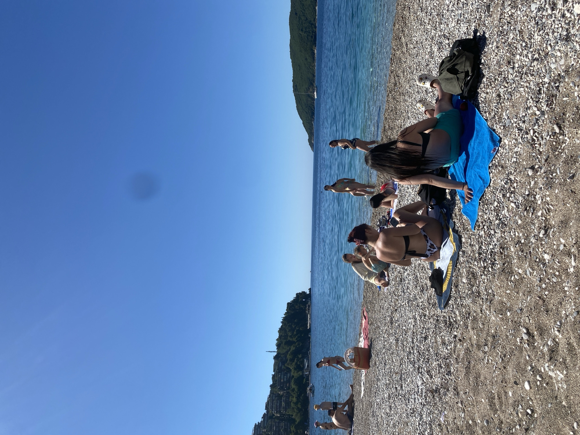 The image shows a pebble beach on a sunny day, with people relaxing and swimming in the turquoise water. The location is near Budva, Montenegro, as indicated by the vegetation and building style on the hillsides in the background. In the foreground, people are sunbathing on towels and sitting on the beach. Some people are standing in the water or swimming. The sky is clear blue.