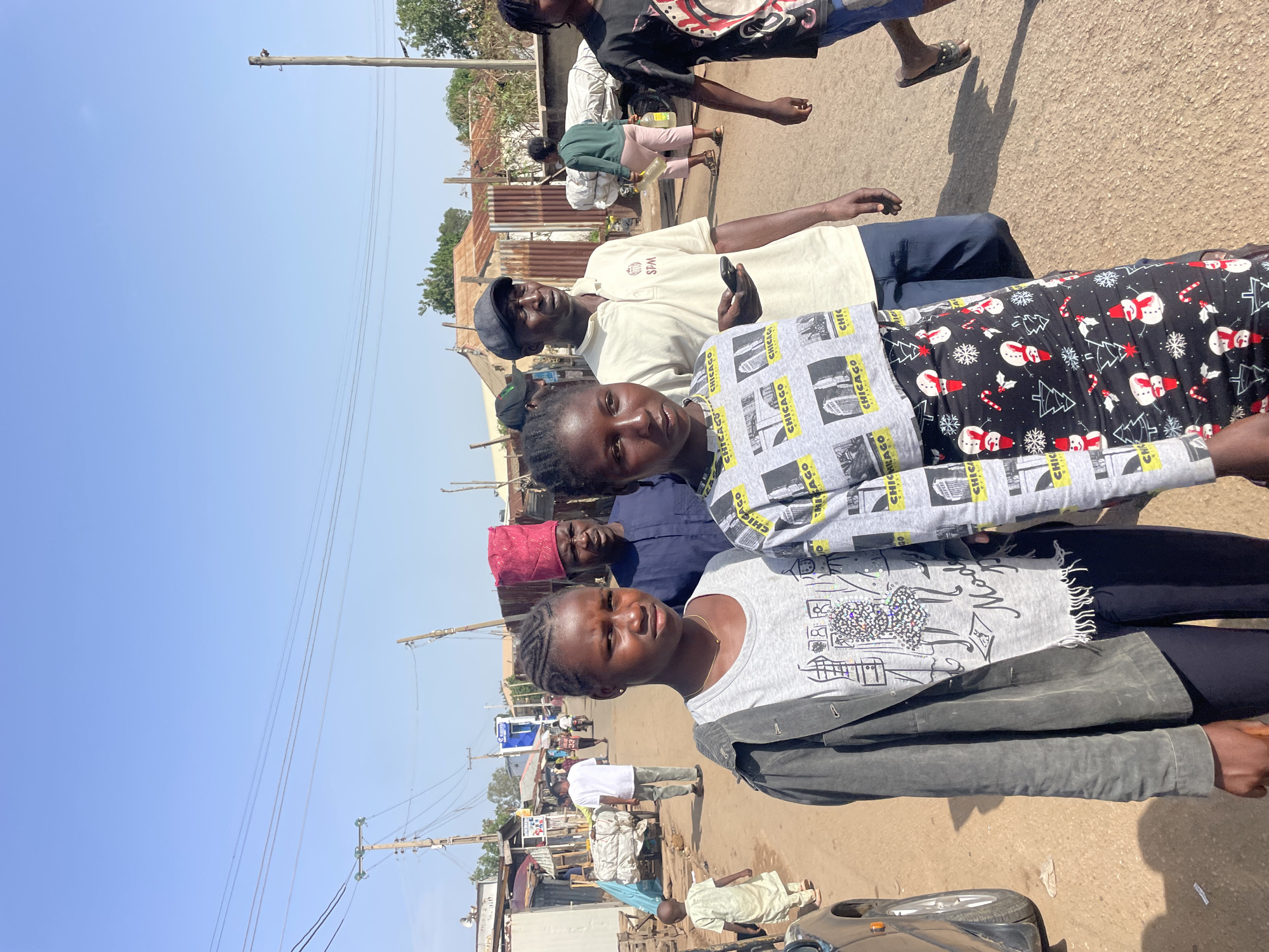 In Gura Top, Nigeria, a group of people stands on a sunlit, unpaved street lined with modest buildings and utility poles. The sky is clear and blue. In the foreground, four people are prominently featured: two younger women in front, one wearing a gray shirt with a black jacket and the other a Chicago-themed sweatshirt, and two older men behind them, one holding a phone. Other people are further down the street, along with parked cars and evidence of market activity.