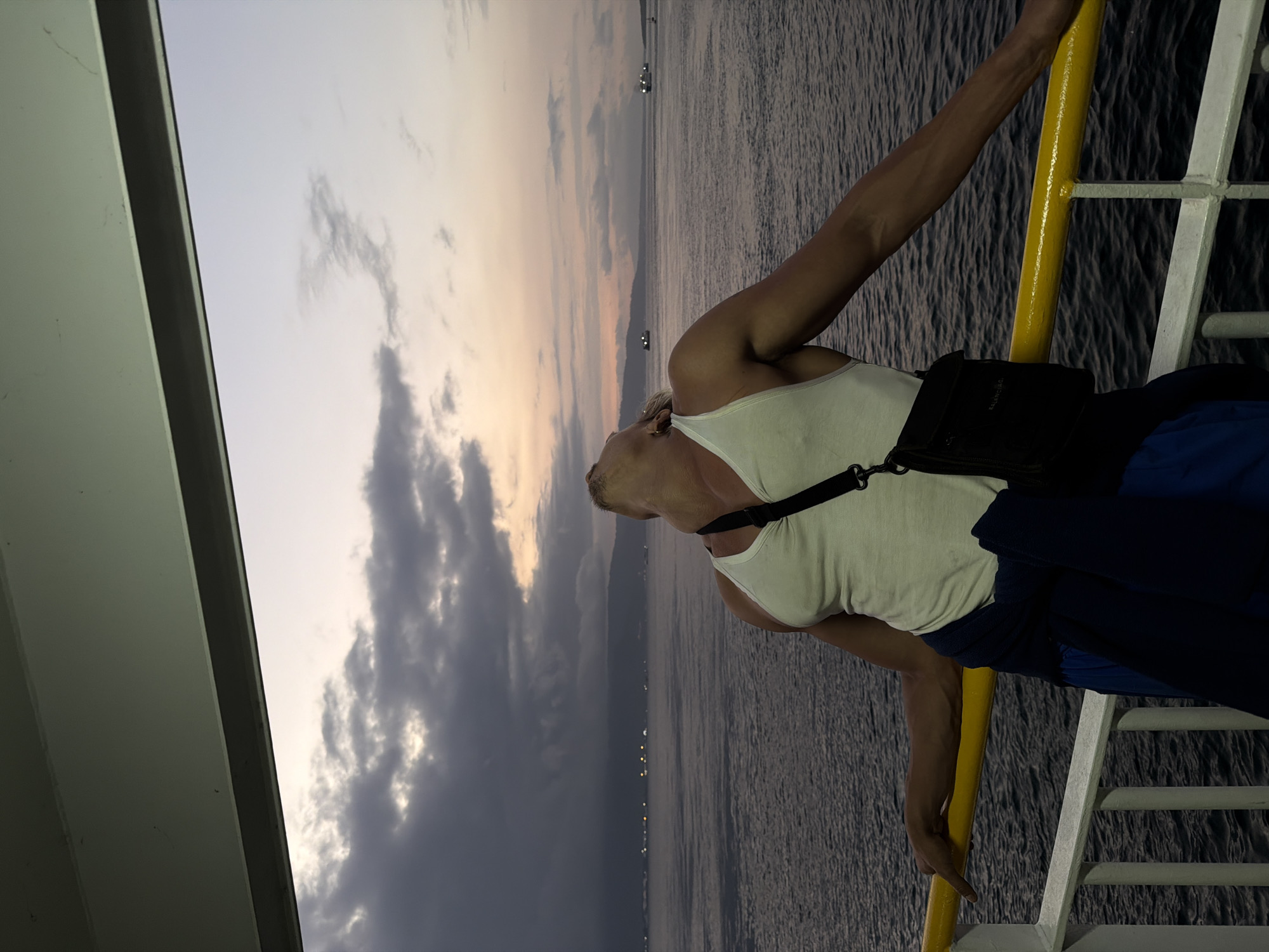 A man with tanned skin, wearing a white tank top and blue shorts, leans back with his head tilted upwards, gazing at the sky from the deck of a boat. A dark crossbody bag is slung over his shoulder, and a dark blue jacket is tied around his waist. He is holding onto a yellow railing on the boat's deck, which overlooks a body of water. The sea is calm, with distant lights visible on a coastline that stretches across the horizon. The sky above is a mix of dark clouds and soft, pastel hues of pink and orange, indicative of either sunrise or sunset. Several boats are visible in the distance on the water. The foreground of the image shows the railing and the deck of the boat, with a portion of a white railing also visible. The overall scene is set at sea, likely near Melaya, Indonesia, given the geographical context provided.