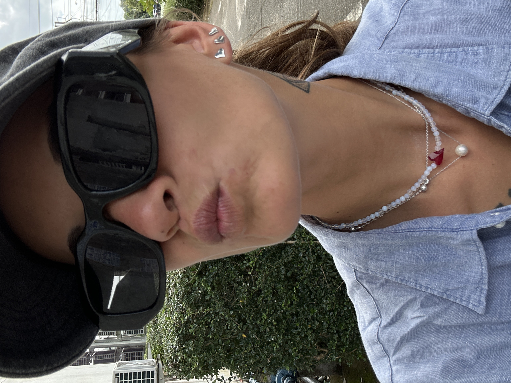 A woman in a blue collared shirt and a dark baseball cap wears large, dark sunglasses and has her lips pursed, as if blowing a kiss. She has three small, silver heart-shaped earrings in her left ear and a tattoo on her neck. She is wearing two necklaces; one with a red heart-shaped pendant and a pearl, and another with white beads. The background shows a dense green hedge, a white building with air conditioning units, and a paved road. The scene appears to be outdoors in a tropical or sub-tropical environment. The overall impression is a candid, casual snapshot.