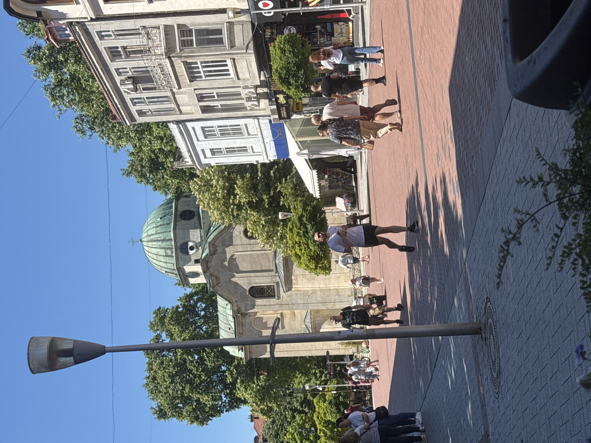 The image depicts a sunny day scene on a pedestrian street in Varna, Bulgaria, specifically in front of the Cathedral of the Assumption of the Virgin Mary. In the foreground, a prominent lamp post stands to the left. The street itself is paved with red bricks. Several individuals are walking in different directions, engaged in casual activity.
To the left, a group of people, appearing to be tourists, are walking away from the camera. Further in the background, a couple walks towards the camera, and a man in a white t-shirt and dark shorts is running with a coffee cup in his hand. A child in a pink dress is also visible walking.
The background features the distinctive green dome of the Cathedral of the Assumption of the Virgin Mary, a significant landmark in Varna. Adjacent to the cathedral are several multi-story buildings with traditional architecture, including shops and businesses on the ground floor. The overall atmosphere suggests a vibrant urban environment with a mix of historical and modern elements. The clear blue sky indicates fair weather.