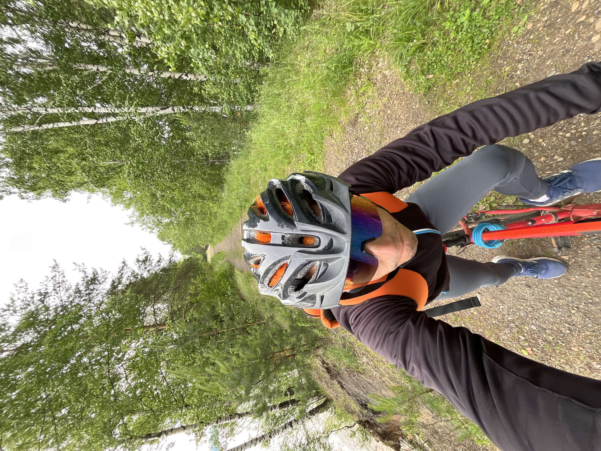 A person wearing a cycling helmet and sunglasses rides a red bicycle on a dirt path next to a body of water and a forest in Chaikovsky, Russia. The person is wearing a black long-sleeved shirt with orange accents, gray cycling pants, and blue athletic shoes. An orange backpack is visible on their back. The path curves ahead, disappearing into the trees. The environment is green and lush with a slightly overcast sky.