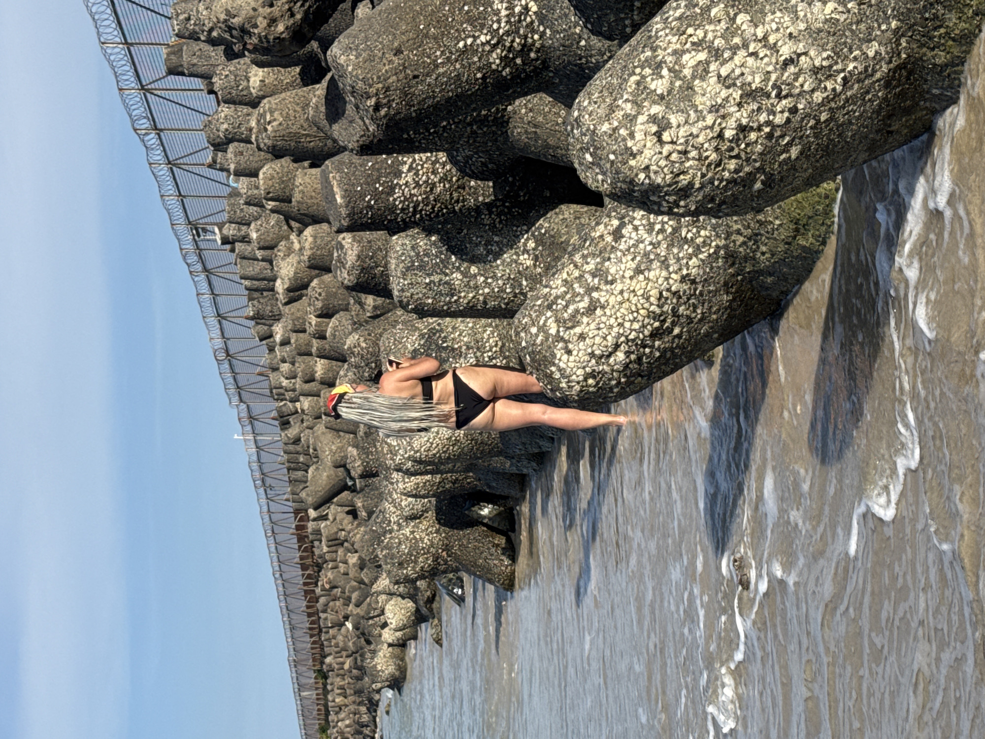 Here is a detailed description of the image:
**Scene:**
The image depicts a scene at Kuta Beach in Bali, Indonesia. This is evidenced by the Tetrapods and the fencing that are commonly found as a coastal defense structure at Kuta Beach.
**People:**
The primary subject is a woman standing near the Tetrapods. She has long, dreadlocked hair, and is wearing a black bikini.
**Objects:**
The dominant feature is the Tetrapod structure. These are concrete structures designed to dissipate wave energy. A fence with razor wire is visible atop the Tetrapods.
**Environment:**
The scene is outdoors, presumably on a sunny day given the brightness of the image. The beach is visible in the background. The ocean water is washing around the base of the Tetrapods. The sky is clear and blue.
**Action:**
The woman is standing near the water's edge, next to the Tetrapods.