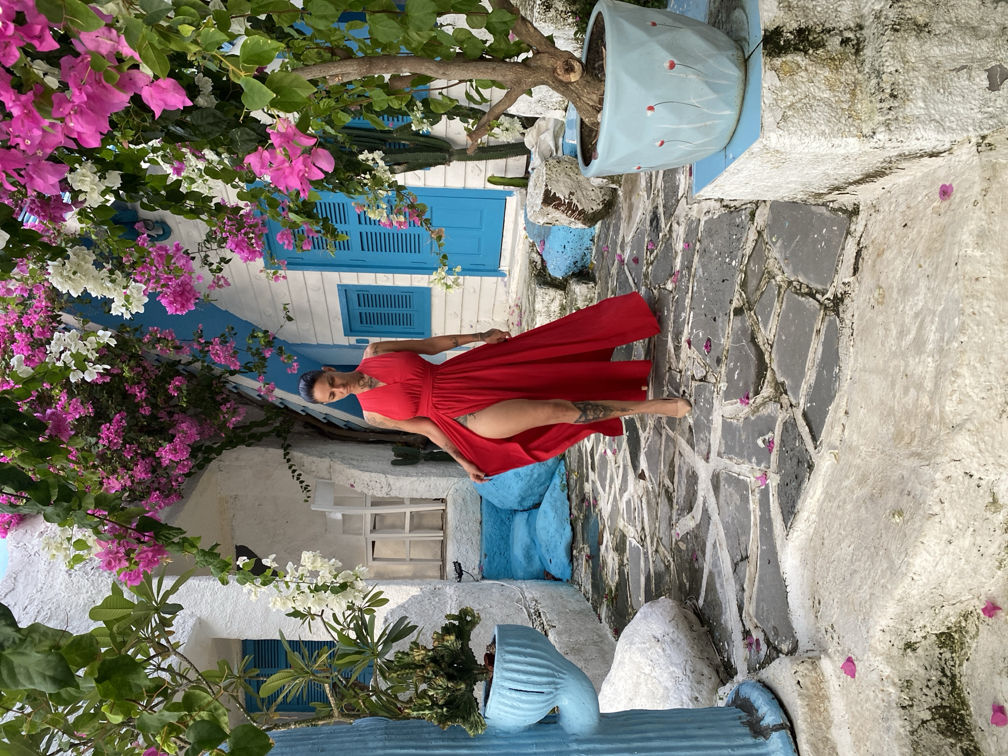 A photograph presents an outdoor scene in Phu Thuy Ward, Vietnam, depicting a woman in a red dress amidst a whitewashed setting with vibrant floral elements. The woman, who has tattoos on her leg and arm, is positioned diagonally in the frame, wearing a v-neck red dress. The surrounding environment features white walls with blue shutters, characteristic of the local architecture. Bougainvillea in shades of pink and white cascade over the buildings, adding a splash of color to the scene. The ground is covered in a gray stone pathway, lightly scattered with fallen flower petals. There are also decorative blue planters containing greenery, complementing the blue accents on the buildings. A cactus is also visible in the background.