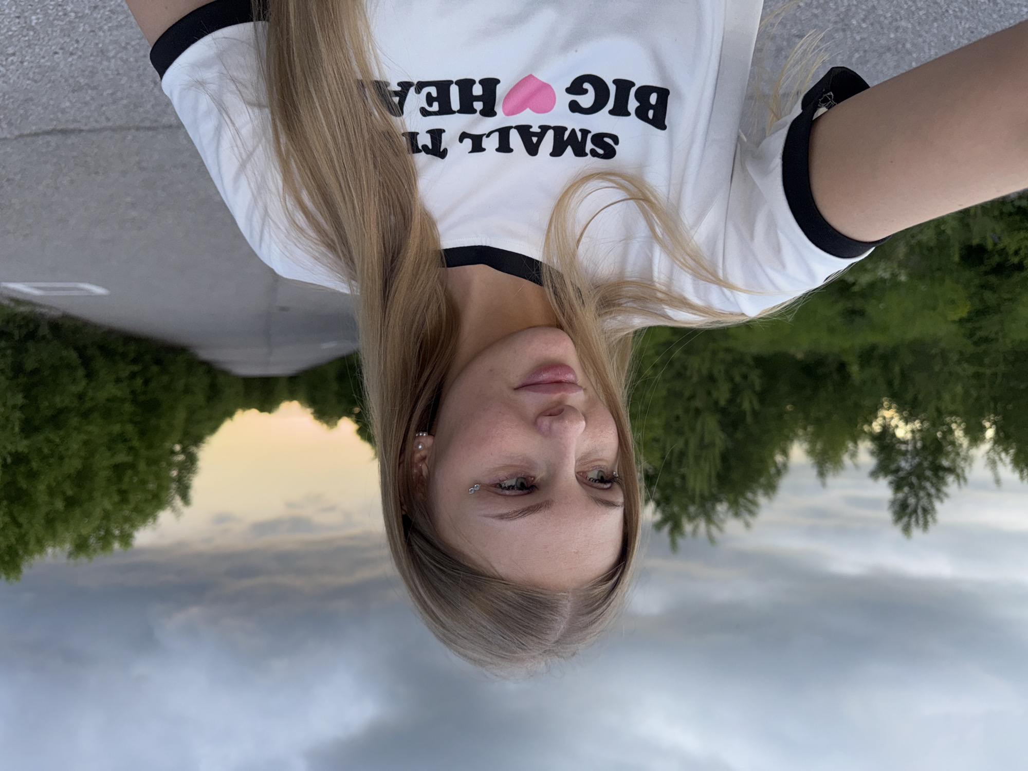 A young woman with blonde hair is captured in a medium shot, looking to her left. She is wearing a white t-shirt with black trim around the collar and sleeves. The t-shirt has black lettering that reads "SMALL TH BIG HEART," with a pink heart replacing the word "HEART." She has subtle eye makeup and a piercing in her left ear. The background consists of a paved road stretching into the distance, flanked by dense green trees on either side. The sky above is overcast with dramatic clouds, transitioning to a lighter, sunset-tinged hue towards the horizon. The scene appears to be outdoors in a somewhat secluded area, possibly a road leading out of a city or through a forested region. The image does not contain enough specific visual information to determine the exact location within the city of Perm, Russia.