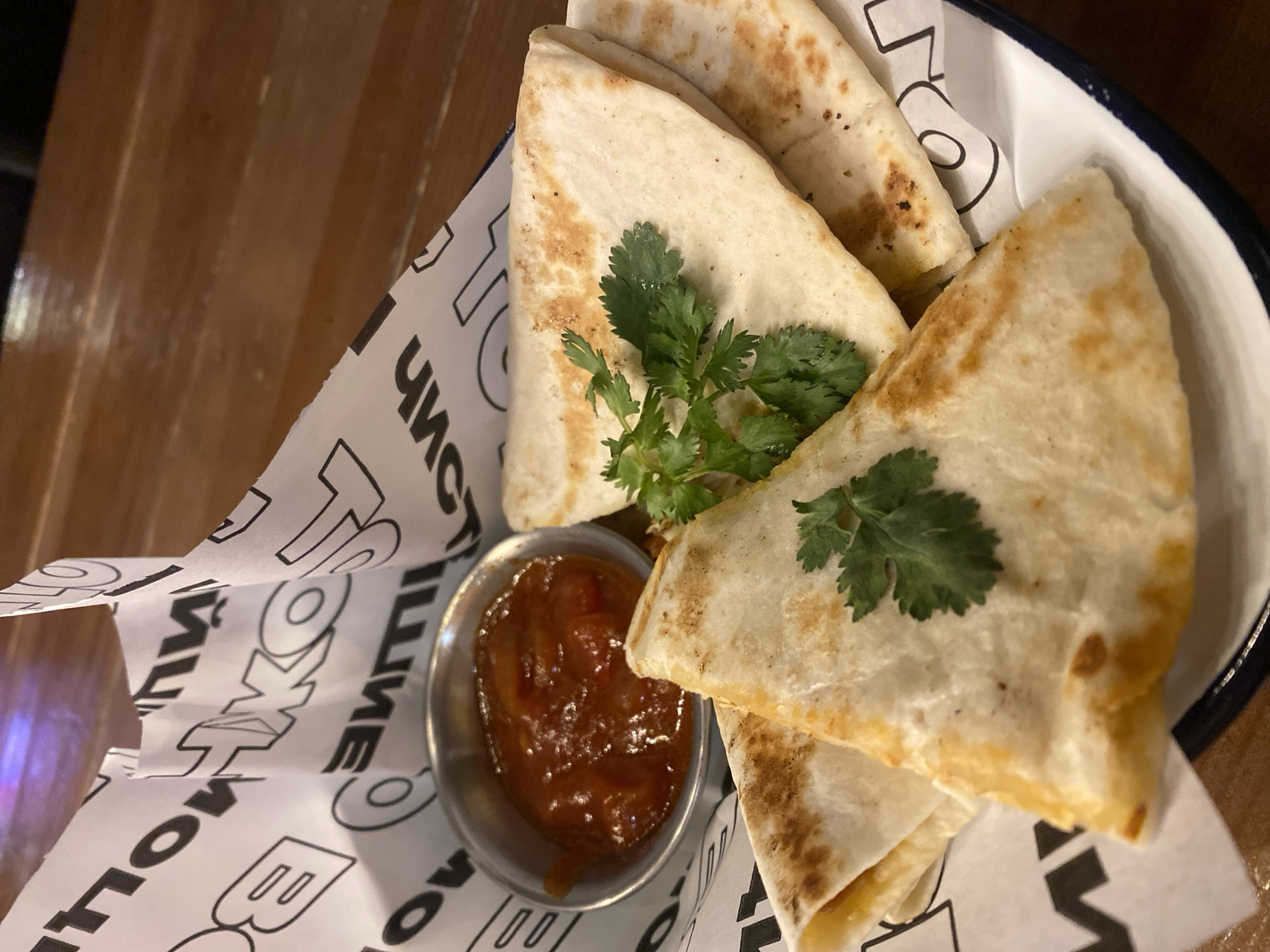 The image shows a metal serving dish filled with quesadilla wedges, garnished with cilantro sprigs, and accompanied by a small metal ramekin of salsa. The dish is lined with printed paper. The letters "TOKYO" and other text in a Cyrillic-like font are visible on the paper. The dish is resting on a wooden surface. The presence of the Cyrillic-like font suggests a theme associated with a cultural representation of Tokyo, although the precise location of the image within Izhevsk, Russia cannot be determined from the visual evidence. There are no people visible in the image. The image focuses on the presented food item and its immediate surroundings.