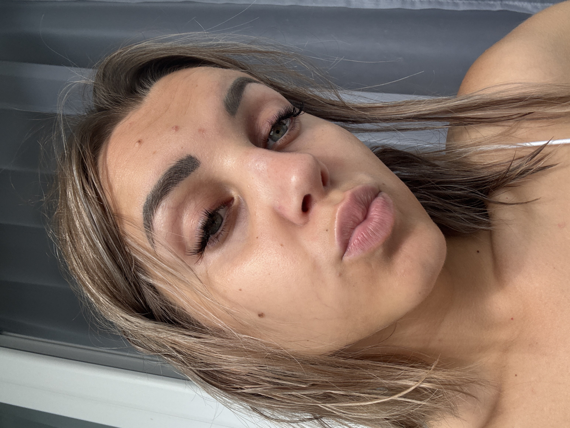 A close-up, selfie-style portrait of a young woman with light brown hair, styled damp and falling around her shoulders. She is facing the camera directly, with her lips slightly pursed. Her eyebrows are dark and well-defined, and she appears to be wearing mascara and false eyelashes. Her skin is fair with some noticeable blemishes on her forehead and cheek. She is wearing a white spaghetti strap top. The background consists of a light-colored wall or curtain to the right and a light-colored vertical surface, possibly a door or window frame, to the left. The image is taken indoors, with soft lighting. There is no indication of Kholmsk, Russia, or any specific city.