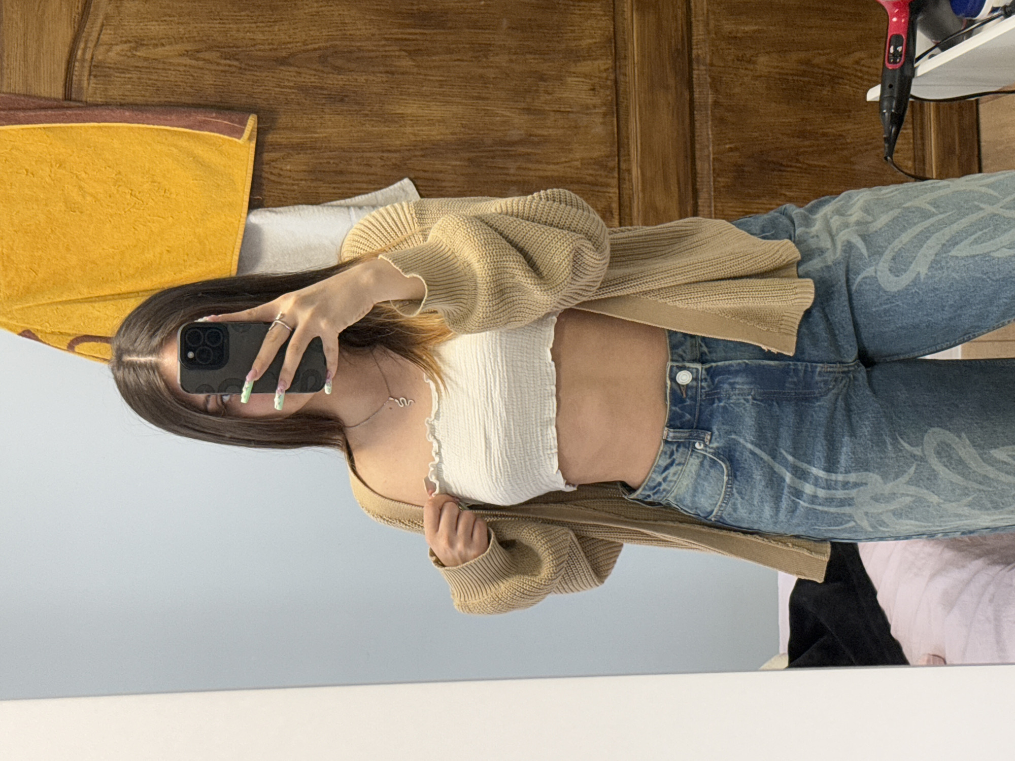 A woman is taking a mirror selfie indoors. She has brown hair and is wearing a white crop top, a beige cardigan, and blue jeans with a white flame pattern. She holds a black smartphone with multiple lenses up to her face. The background includes a wooden door, a yellow towel, a white object, a black object, and a pink bed sheet. The reflection in the mirror also shows a light blue wall and a white surface, suggesting the room she is in. There is no information available to determine the specific location within the city of Sofia, Bulgaria.