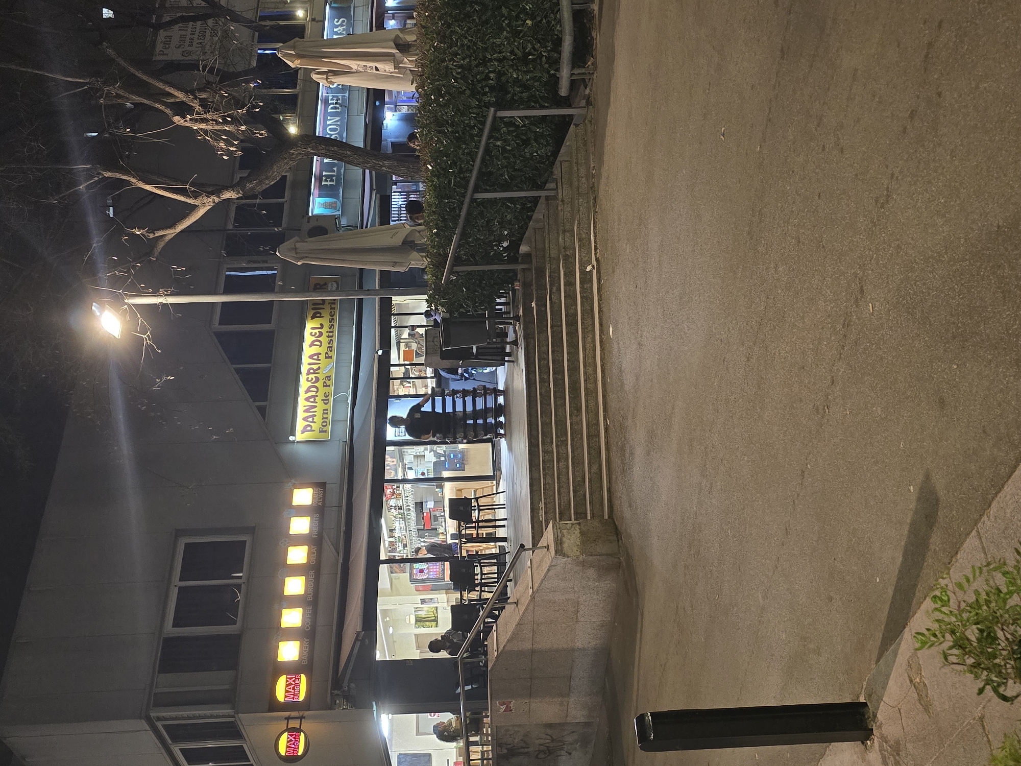 Here's a detailed description of the image:
**Scene Overview:**
The image captures an urban scene at night in what appears to be a commercial area. The scene is centered around a bakery/café named "PANADERIA DEL PILOR" (Forn de Pà Pastisseria). The perspective is slightly rotated.
**Specific Elements:**
* **Bakery/Café:**
* Signage in two languages (Spanish and Catalan).
* The exterior features a set of stairs leading up to the entrance.
* There are patrons inside and in front of the establishment.
* The windows display pastries and other products.
* Above the main sign, there are illuminated signs indicating the offerings: Bakery, Coffee, Burger, Gelato, Fried items ("Fregits"), and "Follastre a l'ast" (likely roast chicken).
* Lower signs for "MAXI Burger" are visible.
* **Exterior:**
* A sidewalk runs along the front of the building.
* There are exterior stairs with metal handrails.
* There are trees with bare branches.
* **People:**
* Patrons are visible inside the café and near the entrance.
* **Environment:**
* It appears to be evening or night, as indicated by the artificial lighting.
* The setting seems to be a moderately busy urban area.
* **Location Details:**
* Based on the signage, we can infer that the location is likely in Catalonia, Spain, given the presence of Catalan. The signs "Peña M San Hd BAR RESTA LA ESQUIAN BERNAC" and "EL SON DES LAS" indicate other commercial establishments nearby.
* Given the bakery's name, it is highly likely that this is located in Cornellà de Llobregat, Spain, named after the neighborhood "El Pilar".