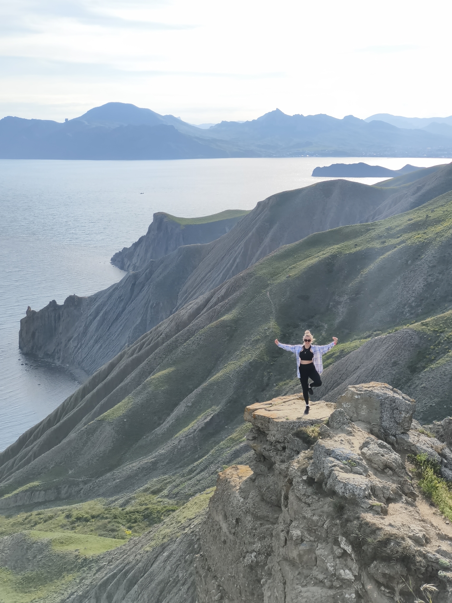 The image shows a person standing on a rocky outcrop in a hilly, coastal region. The person is female-presenting, fair-skinned with blonde hair, wearing a black top and leggings, and a plaid shirt, and is in a balancing yoga pose with arms outstretched. The background features a body of water, likely the Black Sea, and a mountainous coastline. The land is a mix of bare rock and sparse vegetation. The location is likely near Орджоникидзевский поселковый совет, Ukraine, given the characteristic coastal landscape.