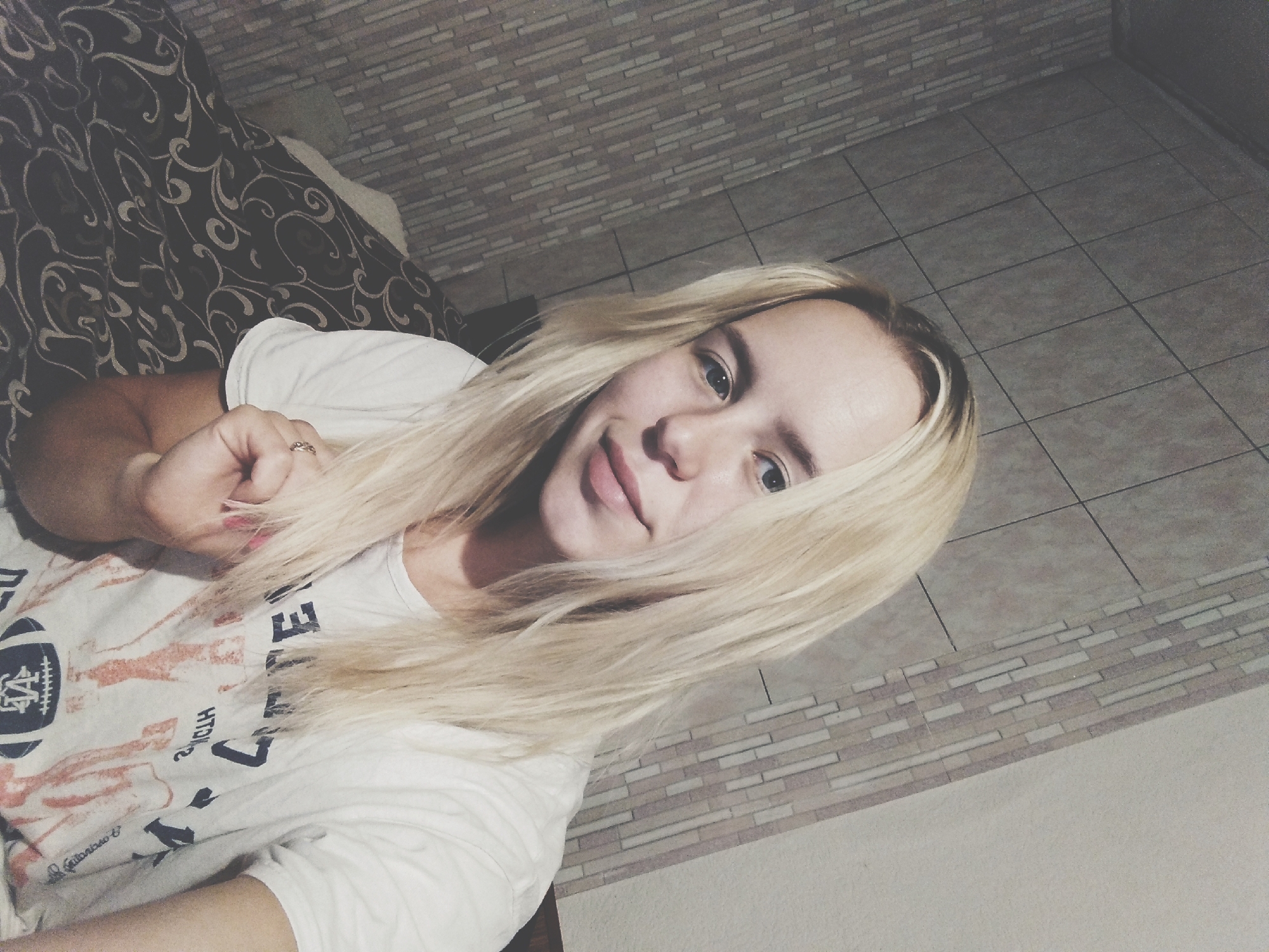 A young woman with long, blonde hair takes a selfie. She is wearing a white t-shirt with graphics on it. Her right hand is raised towards her face and she is wearing a ring. The background appears to be a wall with textured tiles and a patterned bedspread. The lighting suggests an indoor setting, possibly a bedroom. The image does not contain any elements that would allow for an accurate determination of its location within the city of Radisne, Ukraine.