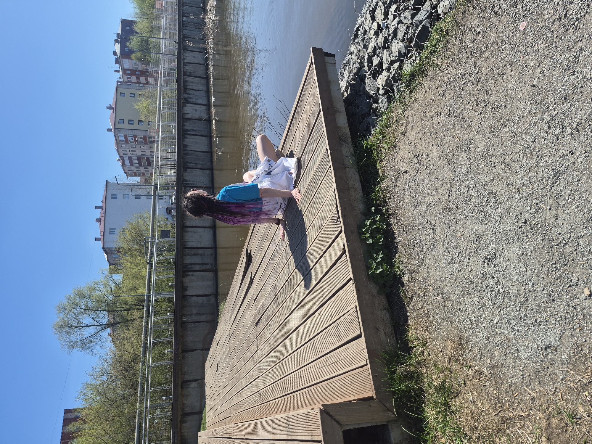 A young person with long, braided hair in shades of purple and brown is reclining on a wooden deck next to a body of water. They are wearing a blue top and white shorts. The deck is adjacent to a pathway covered in gravel, with vegetation growing along the edge. Behind the deck and water is a concrete embankment. Buildings are visible in the distance. The image suggests an outdoor, urban setting, possibly in Votkinsk, Russia.