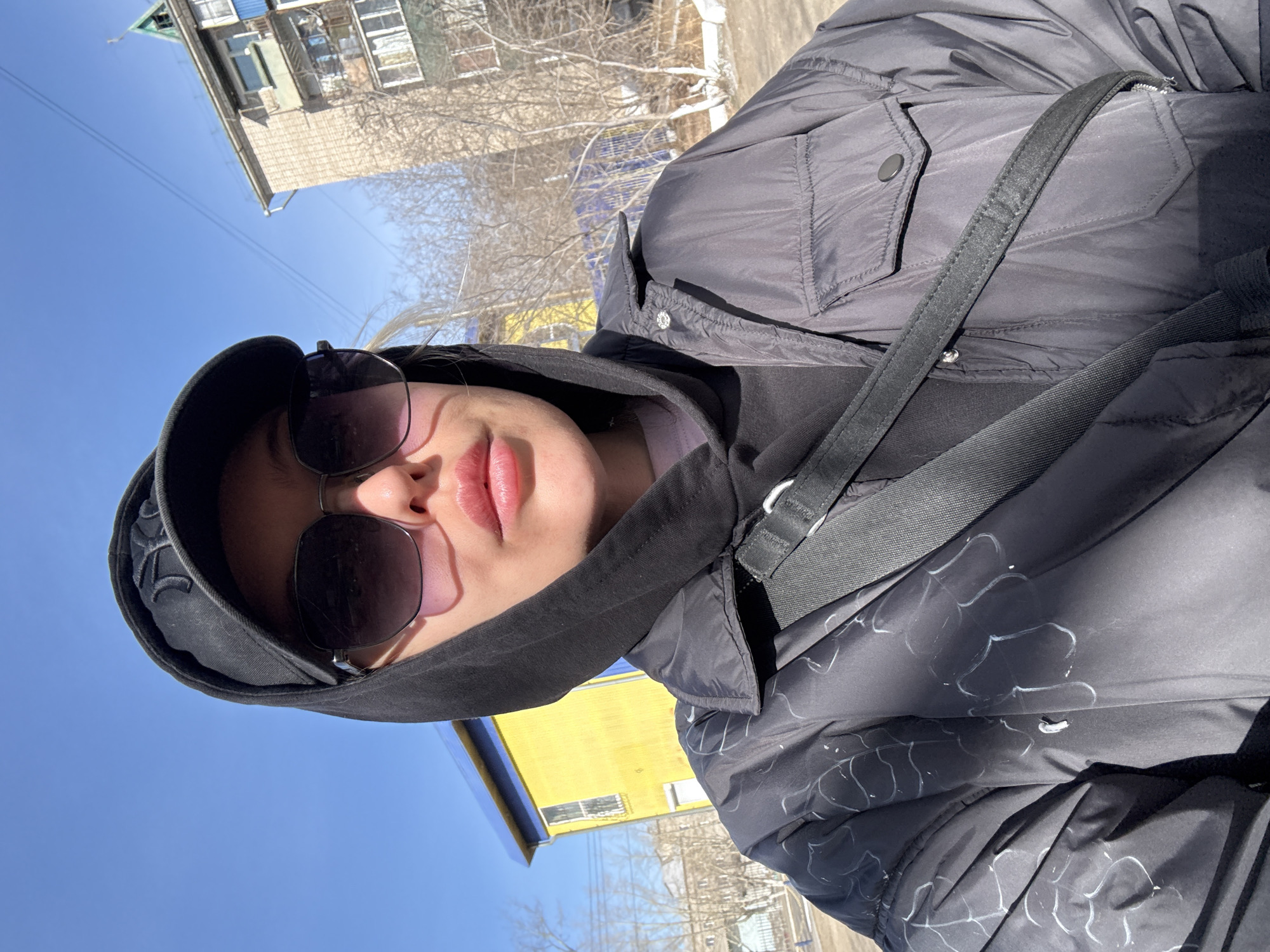 The image is a bright, sunlit outdoor selfie taken from a low angle, looking up at the subject on a clear day. The primary subject is a person, likely female, whose face is partially visible. She wears large, dark sunglasses with a pinkish tint, obscuring her eyes. A black hood covers her head, under which a black baseball cap with a partially visible white embroidered logo (resembling a stylized 'NY') is worn. Her lips are closed, appearing naturally pink, and her expression is neutral.
She is dressed in a black, puffy winter jacket featuring distinctive white, abstract, organic-like patterns across its surface. A black fabric strap, likely from a shoulder bag, crosses her upper body.
The background shows a vibrant, clear blue sky. In the distance, several buildings are visible. One prominent structure has a light-colored facade, multiple windows, and a green-toned roof or trim. Another building in the mid-ground is striking with its bright yellow walls and blue accents. Bare deciduous trees, some with what appears to be a light dusting of snow or frost on their branches, are scattered throughout the background, suggesting late winter or early spring. Thin overhead wires are also noticeable against the sky. The strong sunlight indicates daytime, casting distinct shadows and highlighting the details of the scene. The setting appears to be an urban or suburban environment in Borzya, Russia.