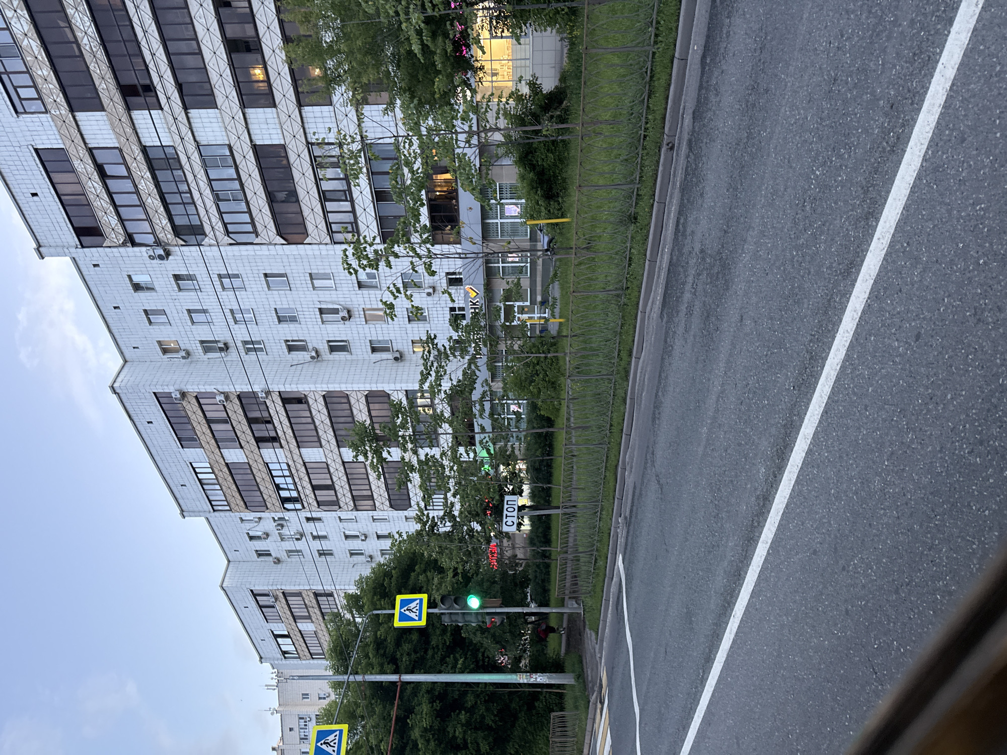 The image shows a street scene in Kazan, Russia, with a modern apartment building and a street with a pedestrian crossing. In the foreground, there is a paved road with a white lane marking. To the right of the road is a sidewalk with a metal fence and green grass behind it. A multi-story apartment building with white tiled facades and dark windows stands prominently on the right side of the frame. On the left side of the image, a traffic light with a green signal is visible, along with a pedestrian crossing sign and a "STOP" sign in Russian. Trees and bushes are planted along the sidewalk and in front of the building. The sky is overcast, suggesting it is either early morning or late evening.
