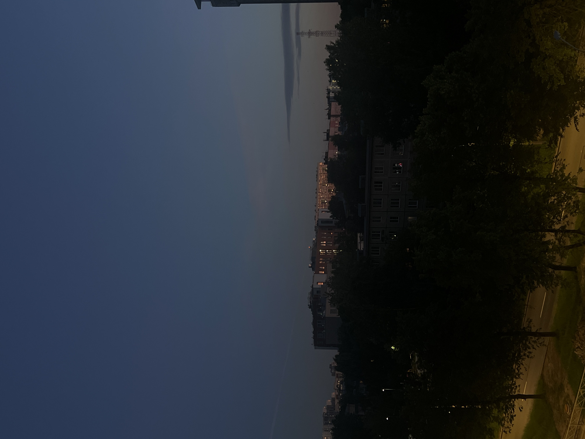 The image depicts a nighttime cityscape in Sinyavino, Russia. The sky is a deep, dark blue, transitioning to a lighter shade towards the horizon. Silhouetted trees dominate the foreground, with a few streetlights casting dim yellow glows. Behind the trees, apartment buildings are visible, their windows illuminated by artificial light, suggesting that residents are inside. In the distance, taller buildings and a transmission tower stand against the twilight sky. The overall atmosphere is one of urban tranquility at the close of day.