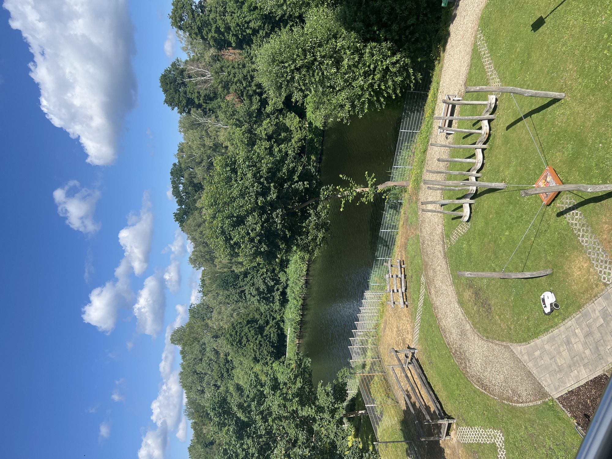 This is an aerial view of a park area next to a body of water, likely the Teltow Canal, in Teltow, Germany. A winding gravel path leads through a grassy area with several playground elements. To the left, there are two wooden benches and a rustic wooden structure. In the center, a row of wooden posts with curved seats attached suggests a balance beam or swing set. In the foreground, a white robotic lawnmower is parked on the grass. Beyond the park, the canal is visible, bordered by lush green trees on the opposite bank. The sky is blue with scattered white clouds.
