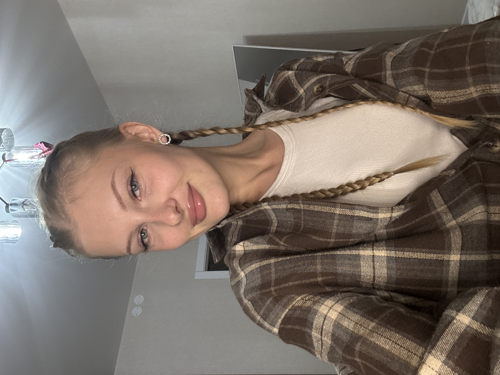The image depicts a young woman with blonde hair in two braids, wearing a brown plaid shirt over a cream-colored tank top. She has on circular, sparkly earrings and appears to be indoors in a room with neutral-colored walls and ceiling lights. There is a mirror visible in the background. The woman is looking directly at the camera with a slight smile. The scene does not contain any specific geographical markers that would place it in Perm, Russia.