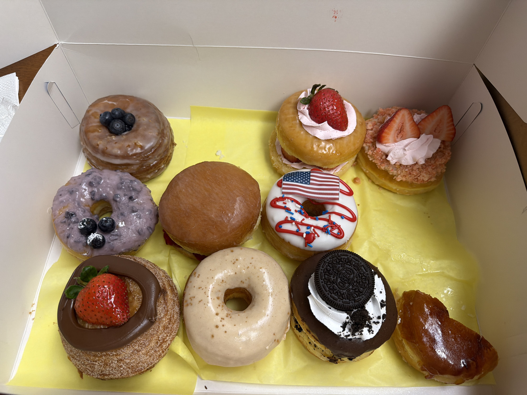 A box of assorted donuts sits on a wooden surface, likely a table. The donuts are arranged in a grid formation on yellow paper. The scene appears to be indoors, with no indication of time of day or weather. There are nine donuts visible:
* A glazed donut topped with blueberries.
* A lavender-frosted donut with blueberries and powdered sugar.
* A plain glazed donut.
* A donut with pink frosting and a strawberry.
* A donut with white frosting, red drizzle, blue sprinkles, and a miniature American flag.
* A donut topped with a strawberry, chocolate frosting, and a leafy garnish.
* A plain beige donut with speckles of cinnamon.
* A chocolate-frosted donut with white frosting, crushed cookies, and an Oreo cookie on top.
* A small, glazed pastry that appears to be a churro bite.
The donuts showcase a variety of colors and toppings, suggesting a celebratory or indulgent occasion. The setting is simple, focusing entirely on the food. No people are visible in the image.