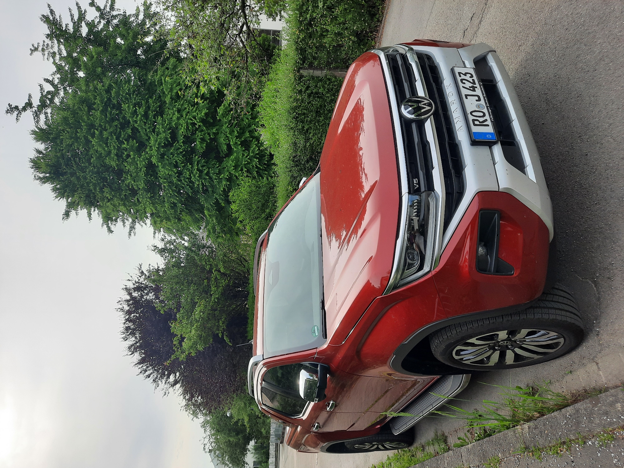 A red Volkswagen Amarok pickup truck is parked on a paved road next to a concrete curb with grass growing out of it. The truck is facing forward and slightly to the right. The truck has a silver trim around the grille and bumper. The license plate is visible and reads "RO J 423". In the background, there are trees and bushes, and a building is partially visible. The sky is overcast. The image was taken in Rosenheim, Germany, as indicated by the license plate and the "Volkswagen Zentrum Rosenheim" text on the license plate frame.